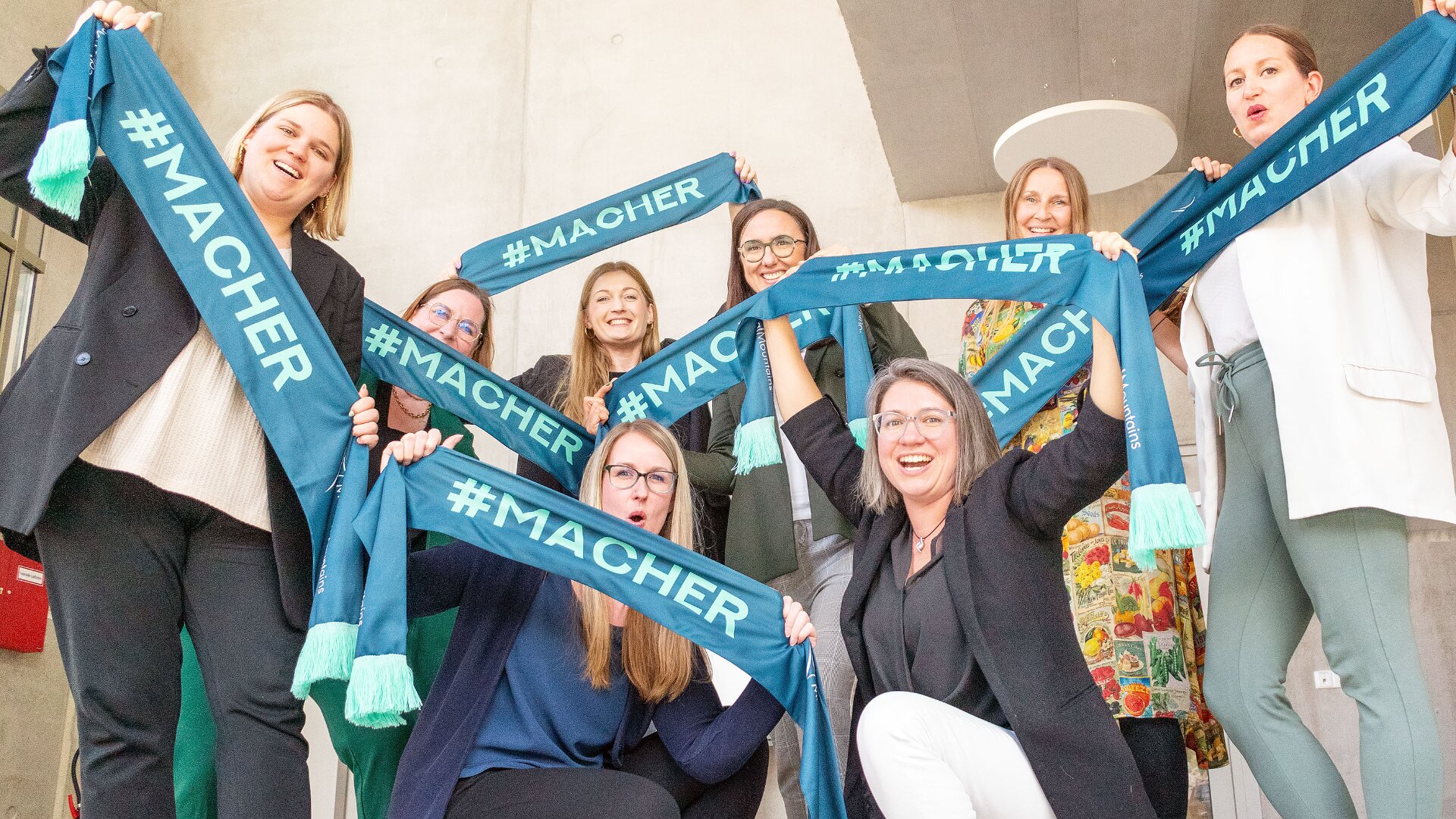 Eine Gruppe von acht lächelnden Frauen posiert in einem Innenbereich und hält begeistert blaugrüne Schals mit der Aufschrift #MACHER in weißen Buchstaben in den Händen. Sie wirken fröhlich und voller Energie und feiern gemeinsam in einem hellen, modernen Raum.