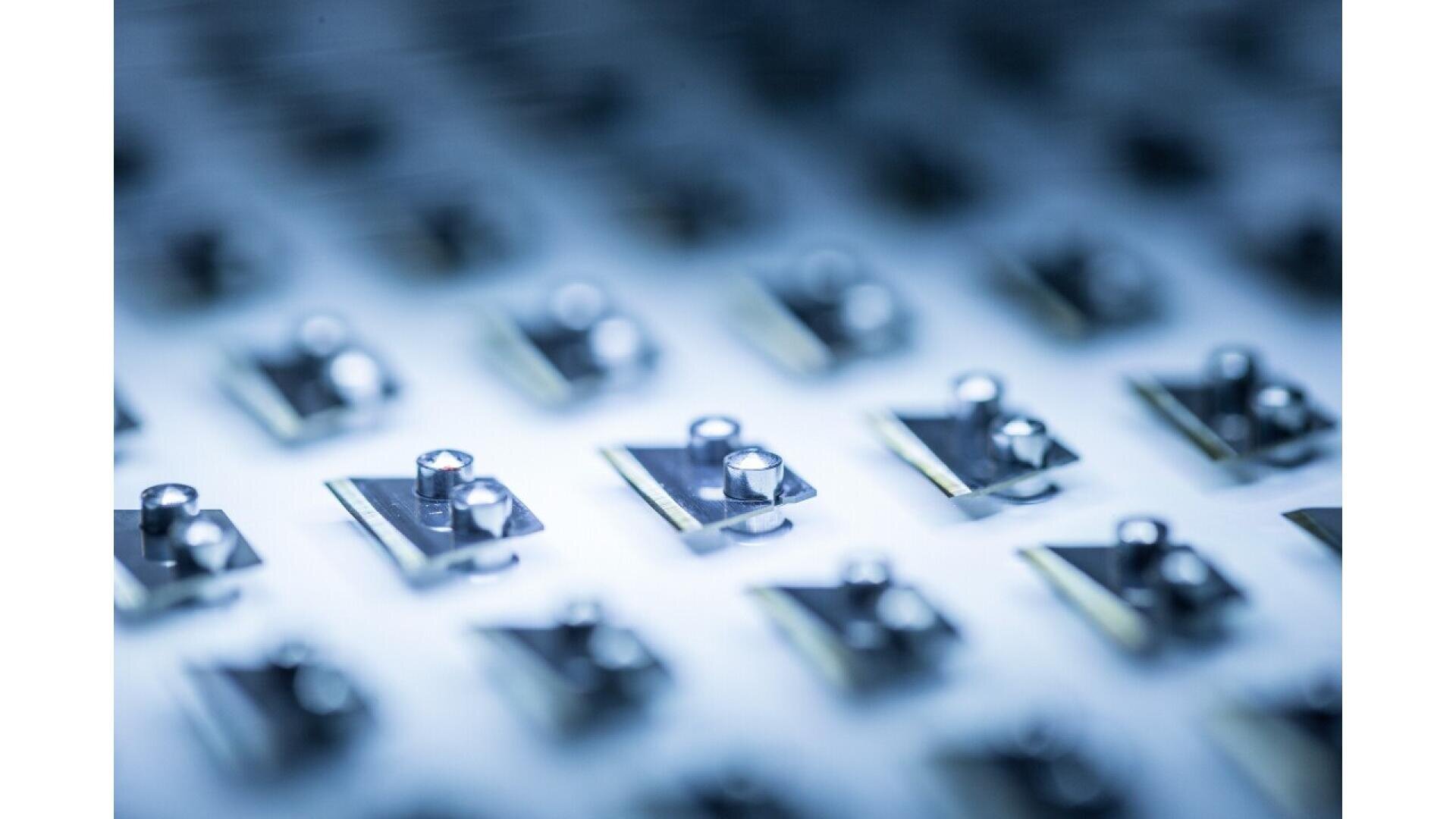 A close-up, macro image of multiple small metallic components arranged in rows on a reflective surface, creating a pattern with a shallow depth of field and a blue-tinted, high-tech appearance.