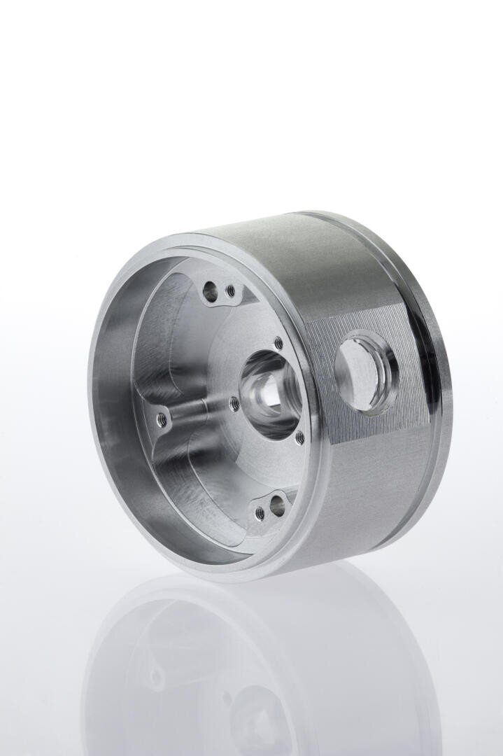 A close-up of a precision-machined metal cylindrical component with several holes, a central cavity, and a threaded side port, set against a white background.