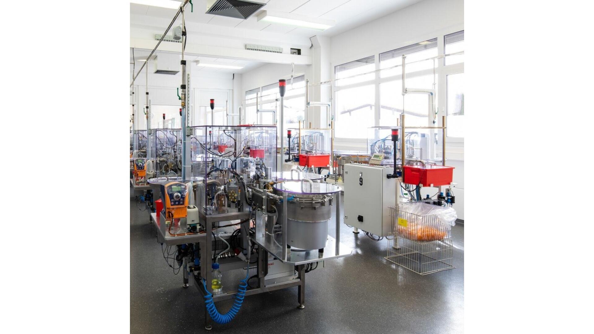 A bright, clean factory room with automated industrial machinery, stainless steel equipment, wires, tubes, and transparent protective shields, positioned near large windows letting in natural light.