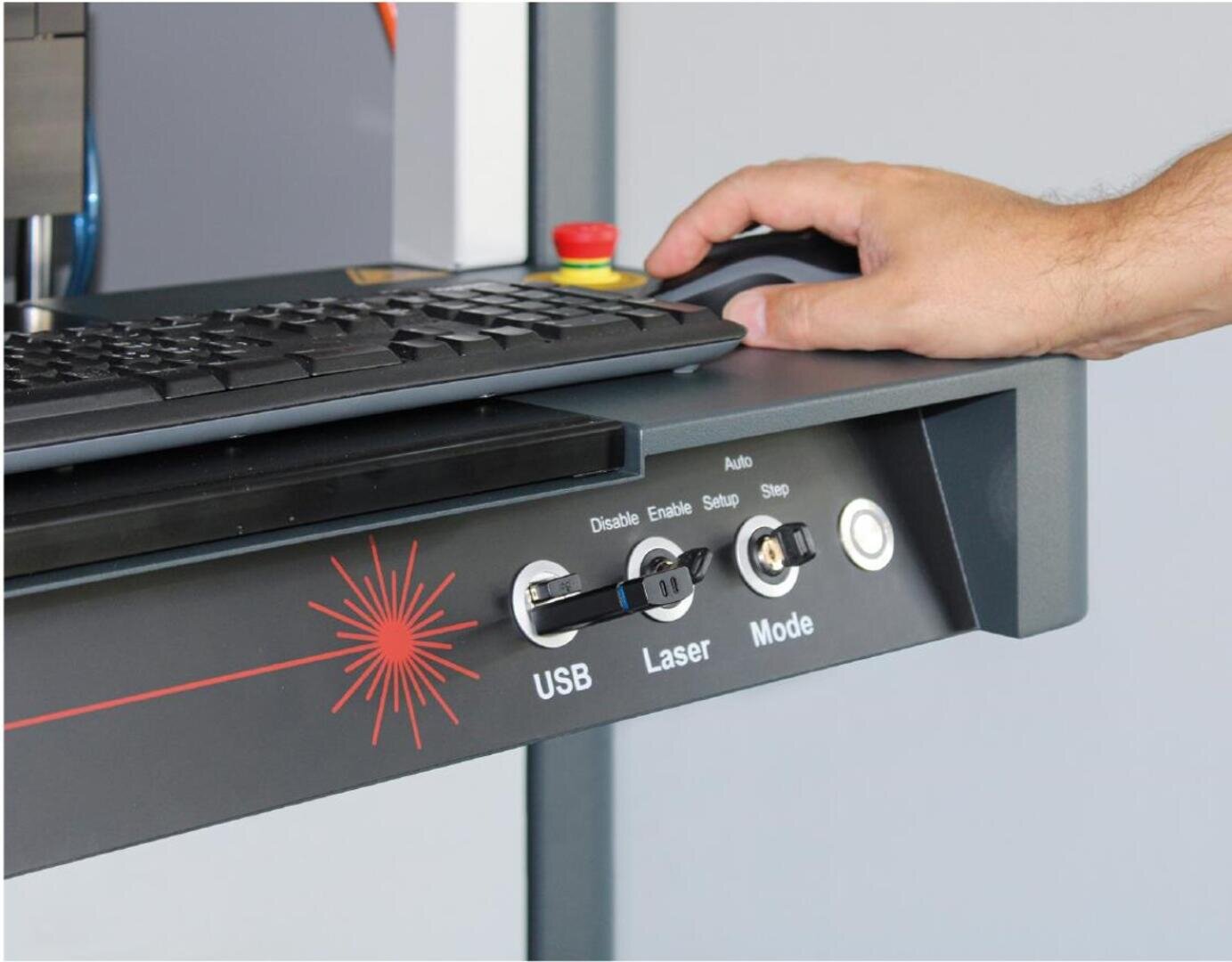 Eine Hand bedient eine Computermaus an einem Arbeitsplatz mit einer Tastatur und einem Bedienfeld, das mit USB, Laser und Modus beschriftet ist und über Schalter, eine Taste und eine rote Not-Aus-Taste verfügt.