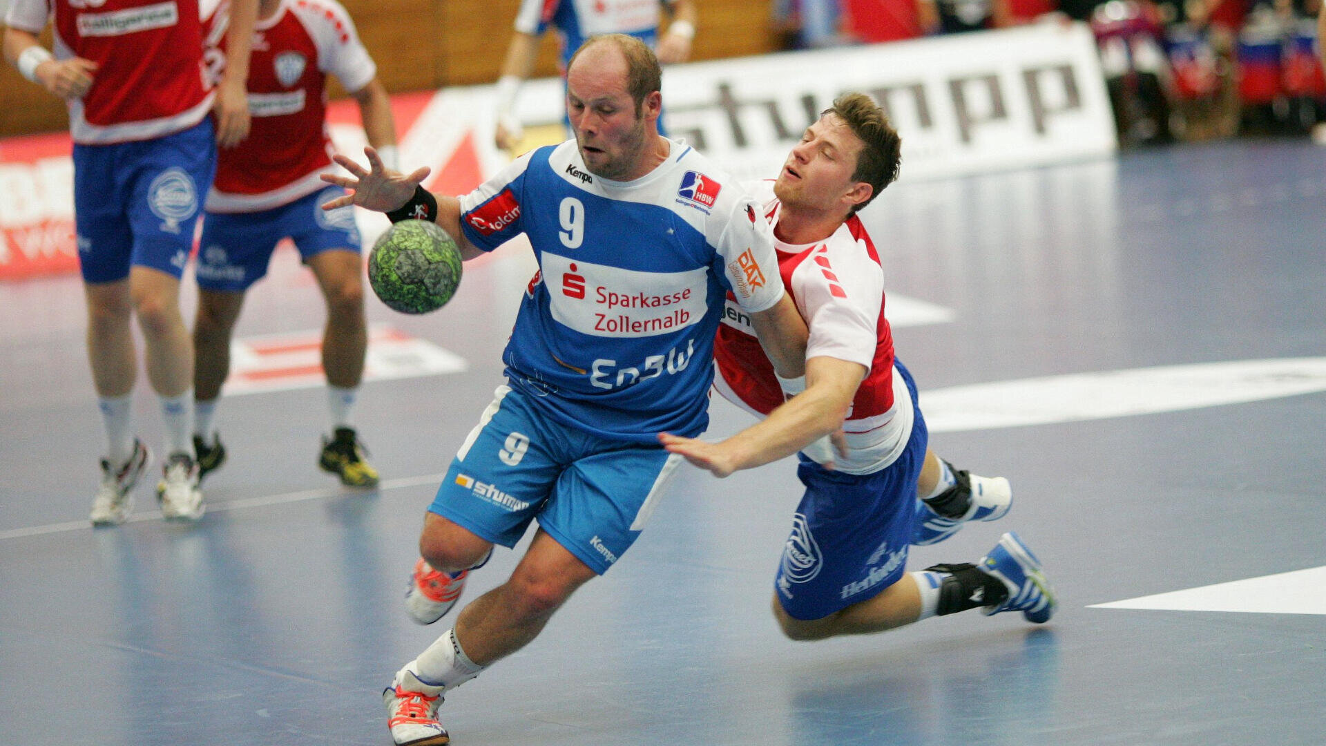 Two male handball players compete for the ball; the player in blue is dribbling while the player in white and red tries to stop him by reaching around his waist. Other players and a referee are visible in the background.