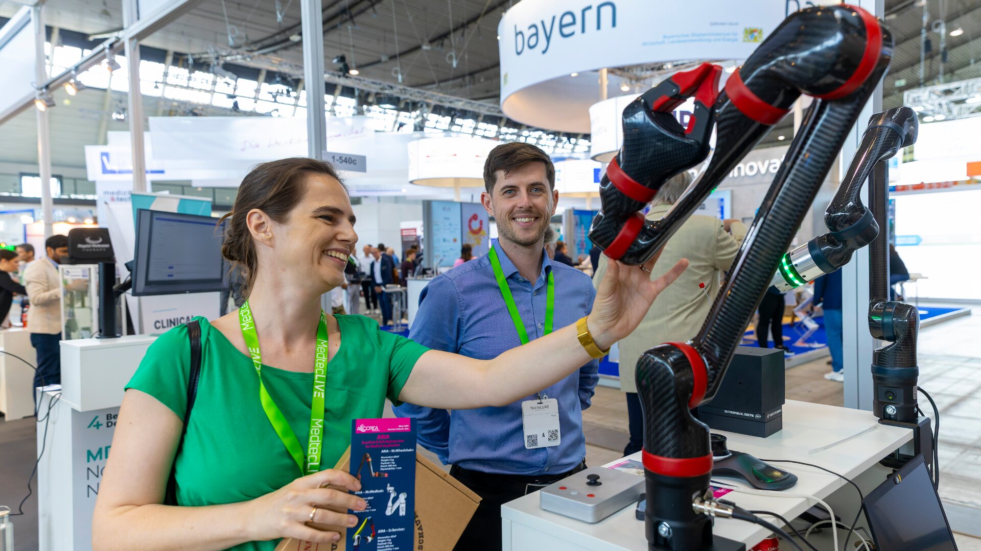 Eine Frau lächelt, während sie auf einer Technologieausstellung mit einem Roboterarm interagiert. Neben ihr steht ein Mann. Beide tragen grüne Schlüsselbänder. Im Hintergrund sind Messestände und Banner zu sehen.