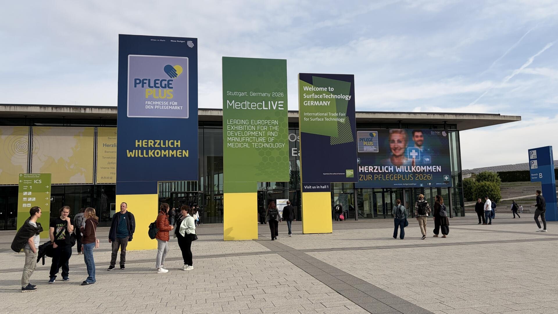 Menschen laufen vor einem Kongresszentrum mit großen bunten Bannern für Veranstaltungen wie Pflege Plus, MedtecLIVE und SurfaceTechnology GERMANY. Auf den Bannern stehen Grußworte und Veranstaltungshinweise in Deutsch und Englisch.