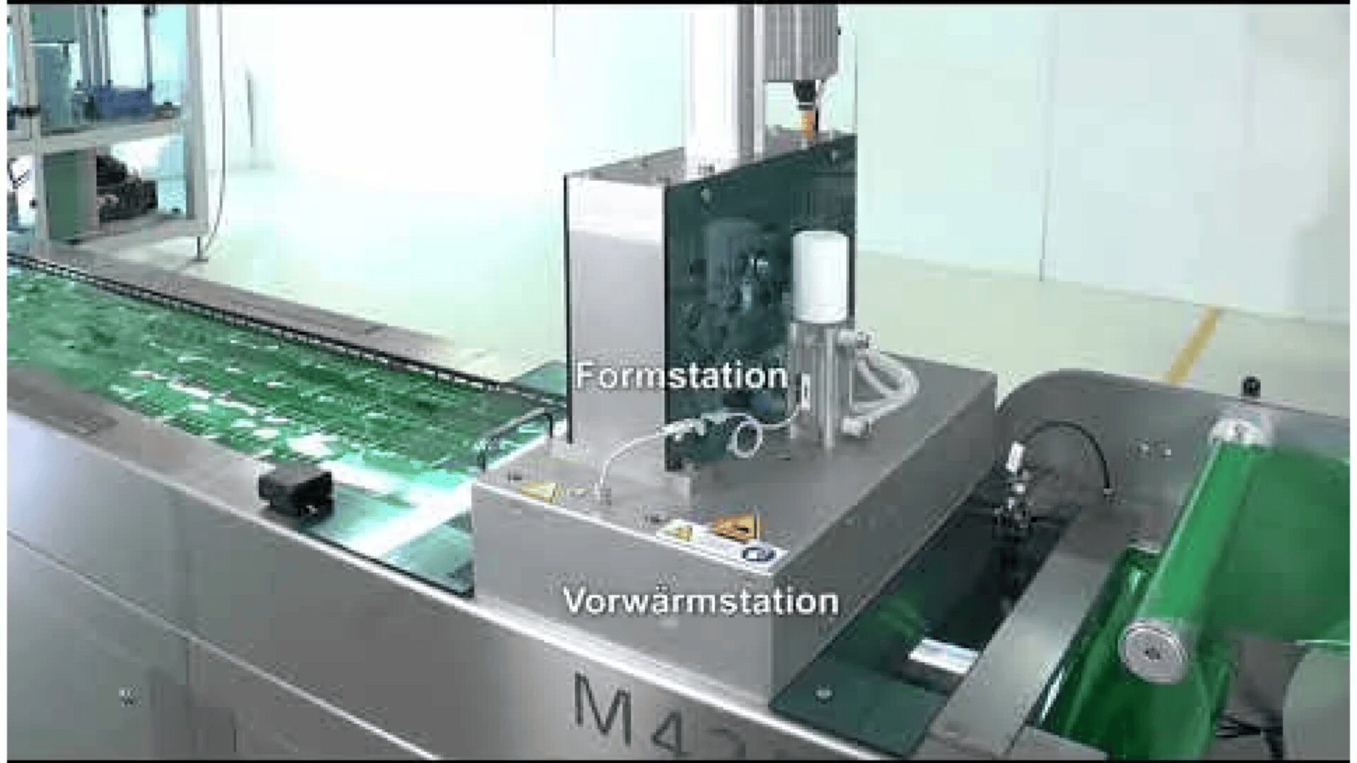 A packaging machine is shown processing green trays, with labels indicating the Formstation and Vorwärmstation (forming and preheating stations in German). The machine has metallic parts and control panels.
