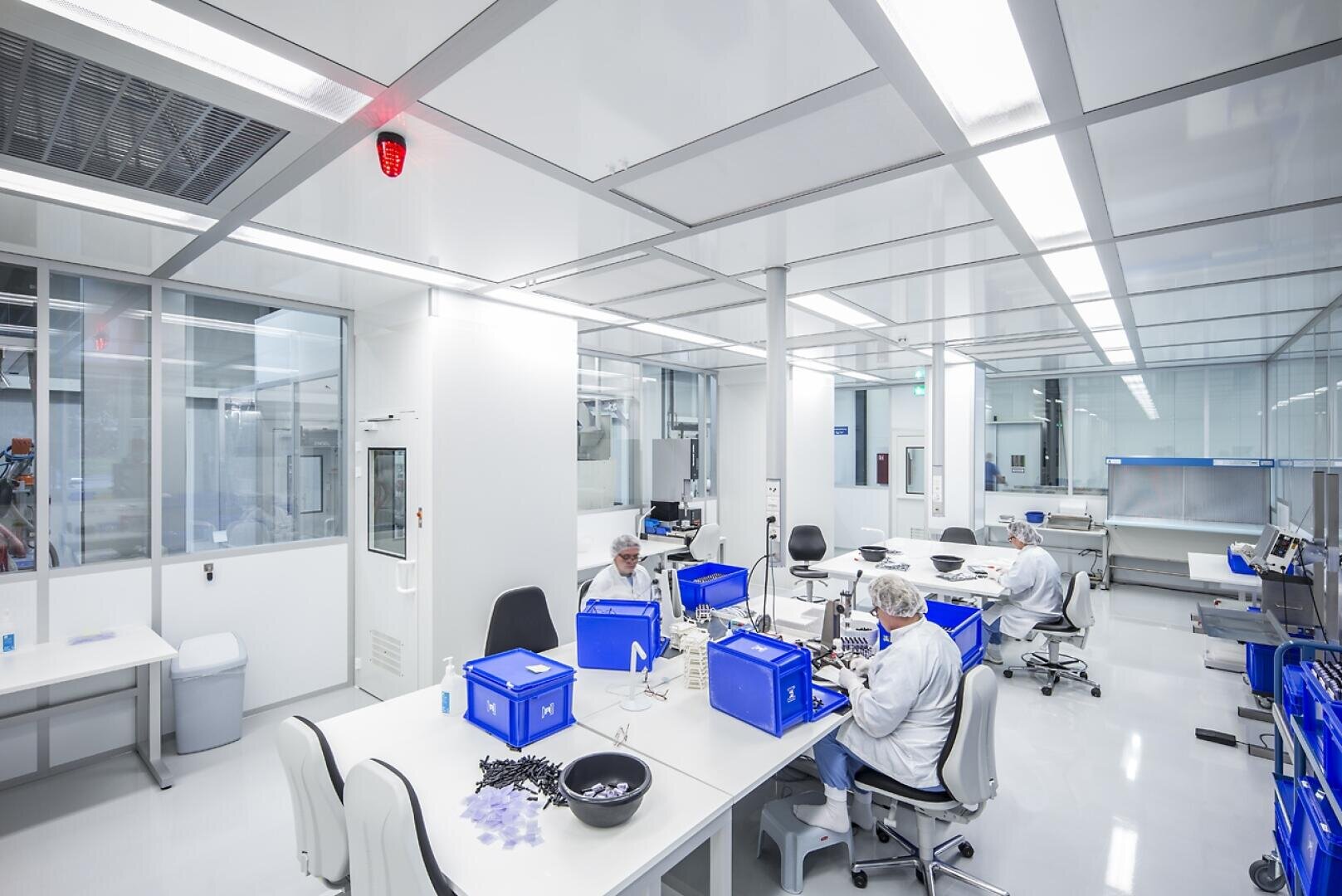 Ein sauberes, modernes Labor mit heller Beleuchtung, weißen Wänden und mehreren Arbeitern in weißen Schutzanzügen und Haarnetzen, die an Tischen mit blauen Lagerbehältern Gegenstände zusammenbauen.