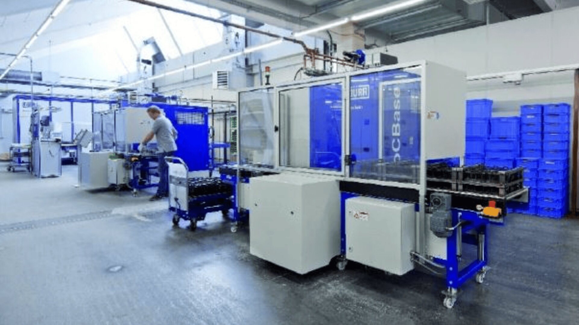 Ein Arbeiter bedient Maschinen in einer sauberen, gut beleuchteten Fabrik. Überall sind blaue Geräte und Plastikkisten gestapelt. Die Umgebung wirkt organisiert und industriell.
