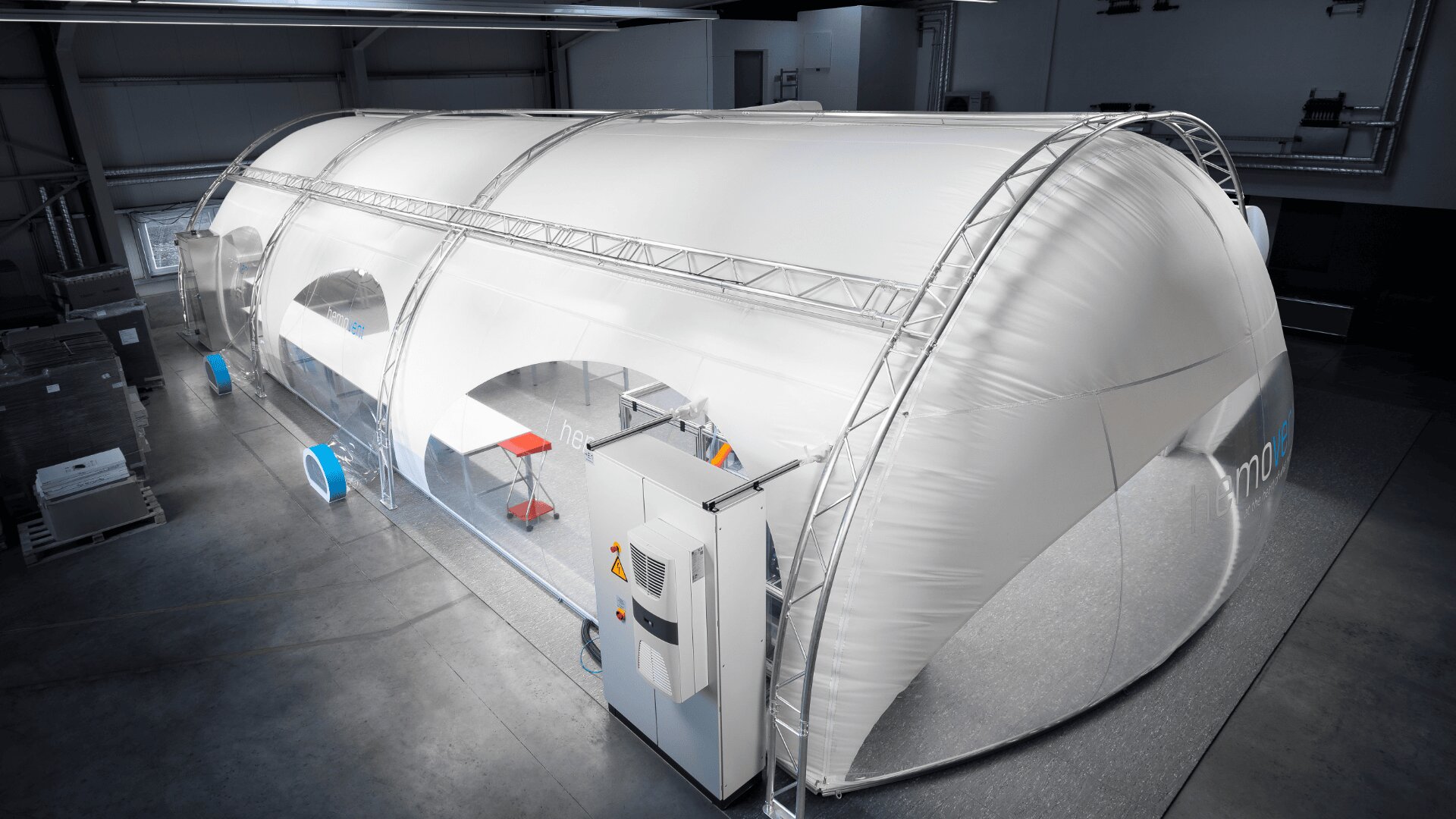 Eine große, weiße, aufblasbare Struktur mit transparenten Teilen steht in einer geräumigen, industriellen Lagerhalle. Tische, Stühle und Geräte sind im Inneren des Gebäudes zu sehen.