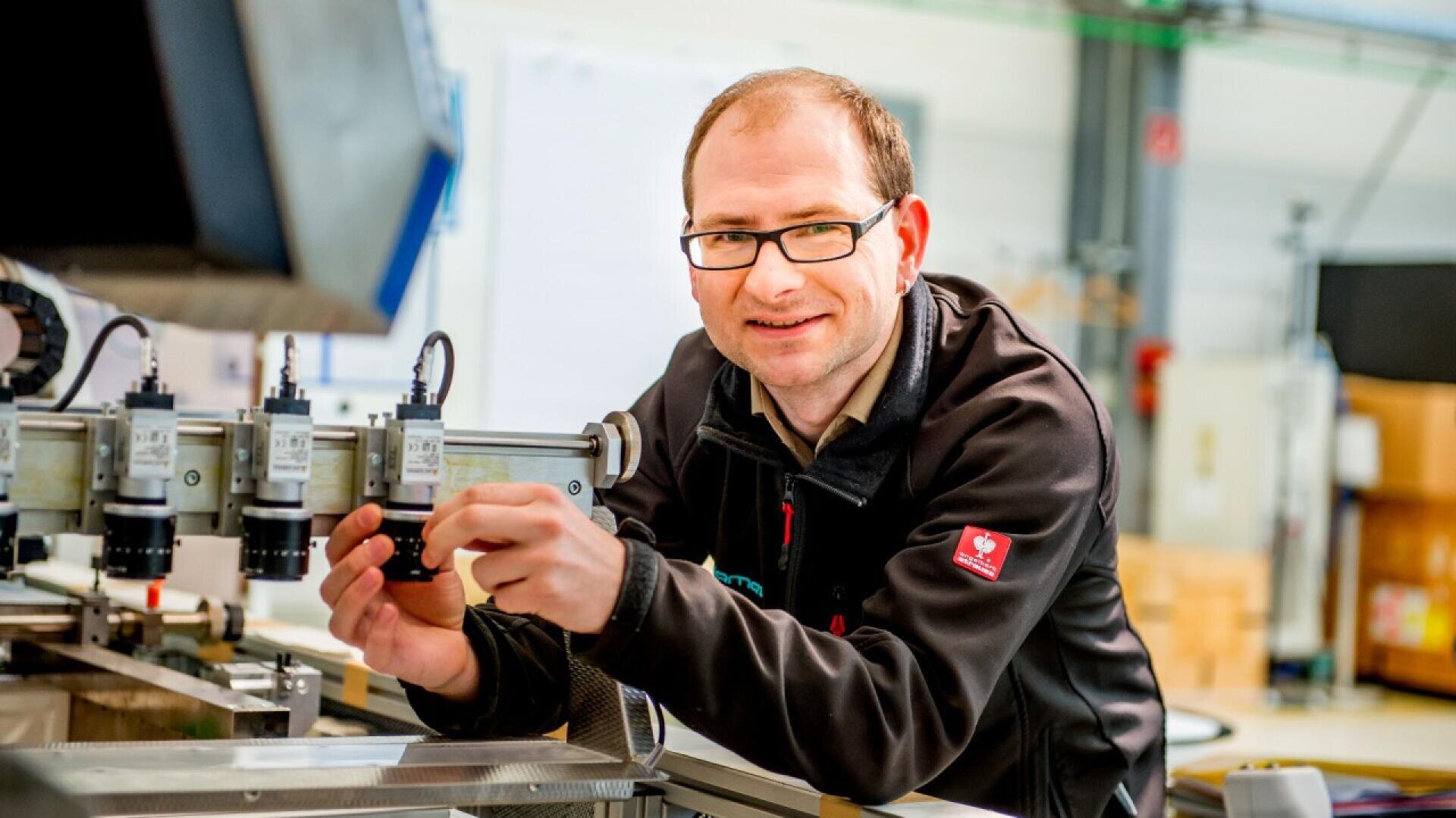 A man wearing glasses and a black jacket smiles while adjusting equipment in an industrial or laboratory setting. Machinery and blurred background elements are visible around him.