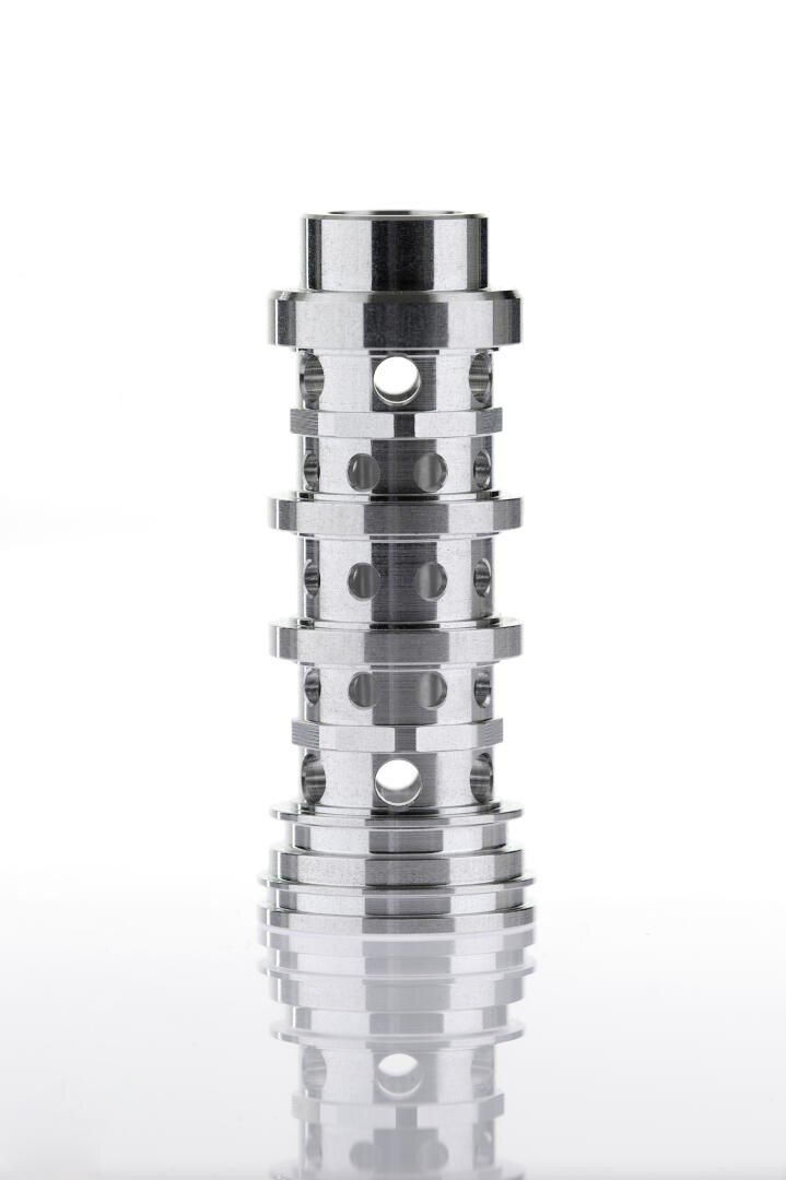 A polished metal cylindrical component with multiple holes and grooves, standing upright on a reflective white surface against a white background.