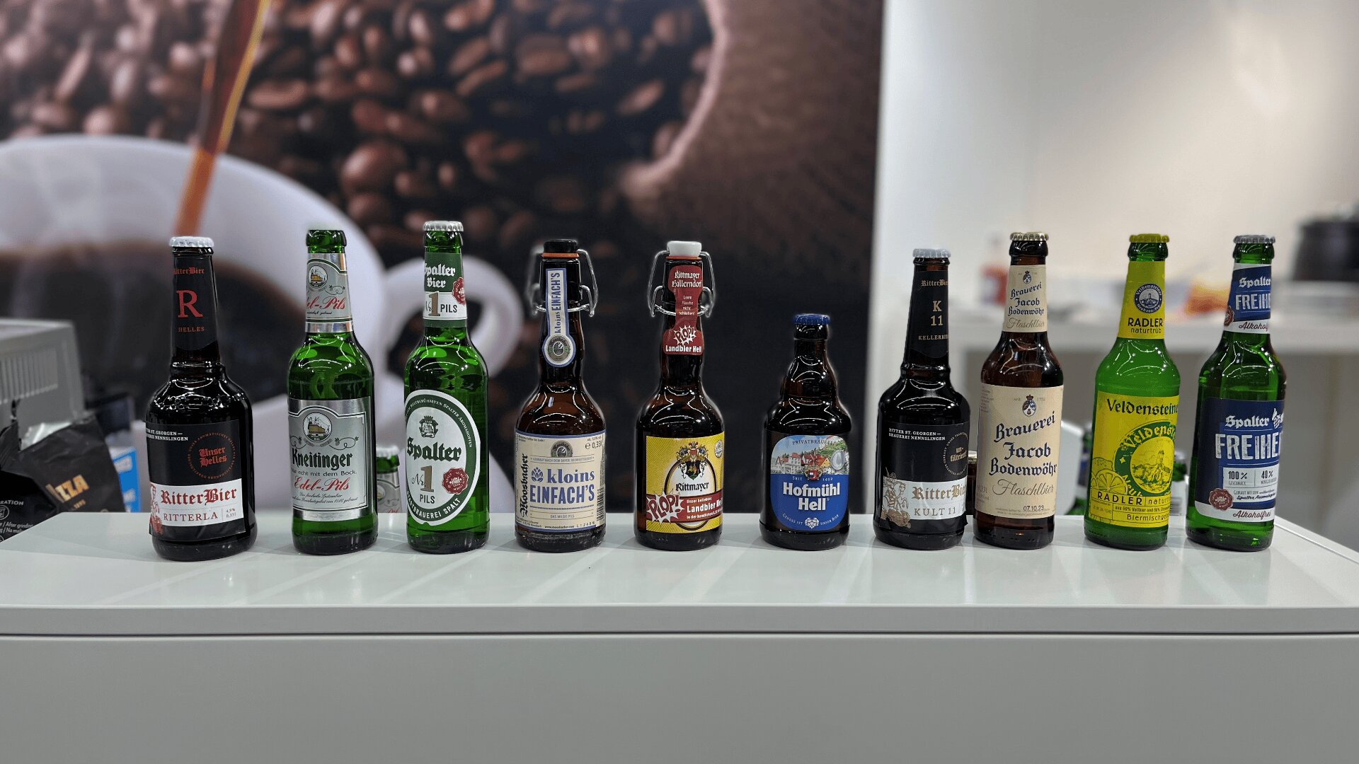 Auf einer weißen Theke vor einem unscharfen Hintergrund mit Kaffeemotiv sind zehn verschiedene Flaschenbiere mit unterschiedlichen Etiketten aufgereiht.