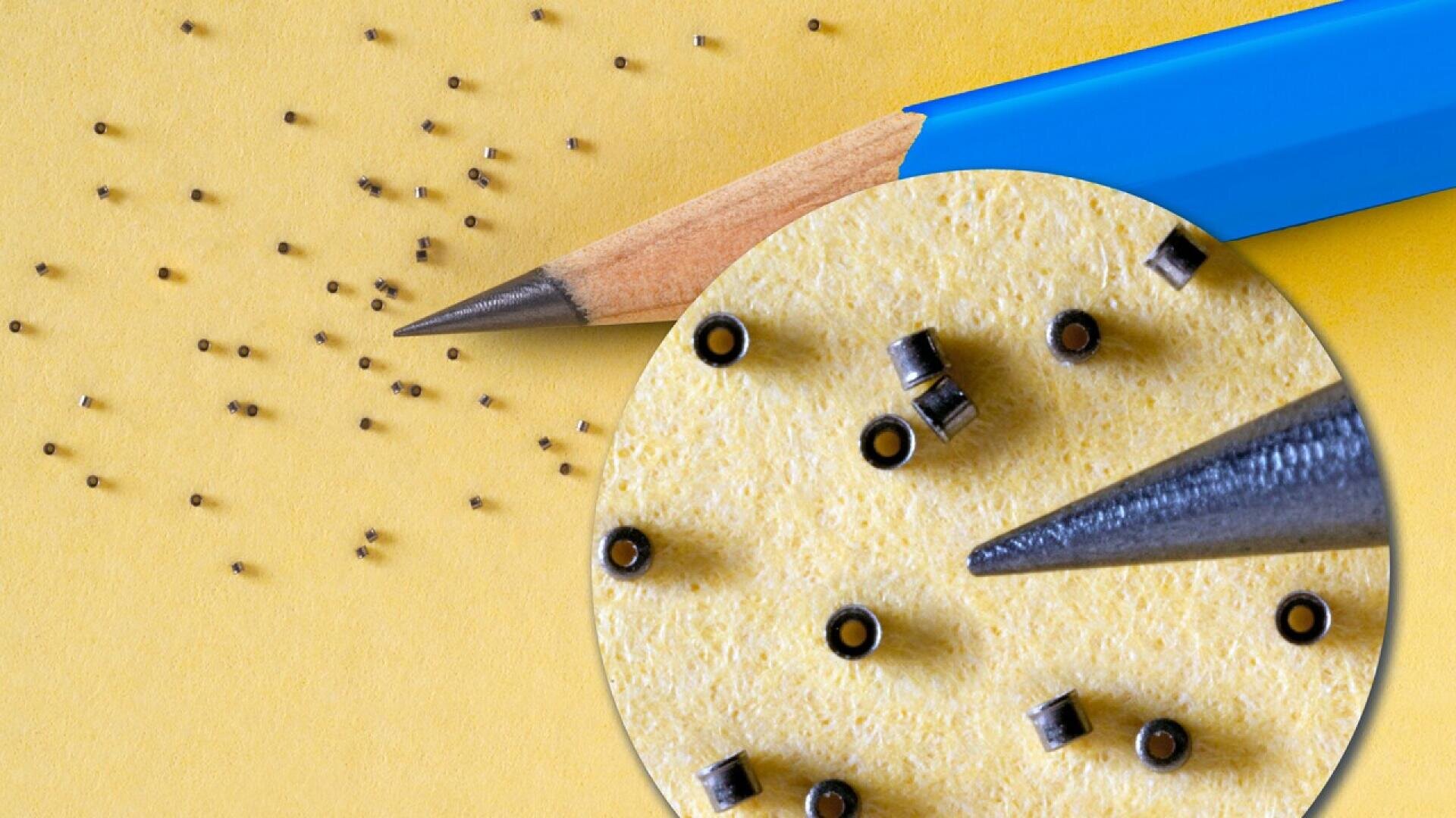 A sharpened pencil on yellow paper next to many tiny black cylindrical objects. A magnified circle shows the pencil tip pointing at the small objects, highlighting their size and detail.