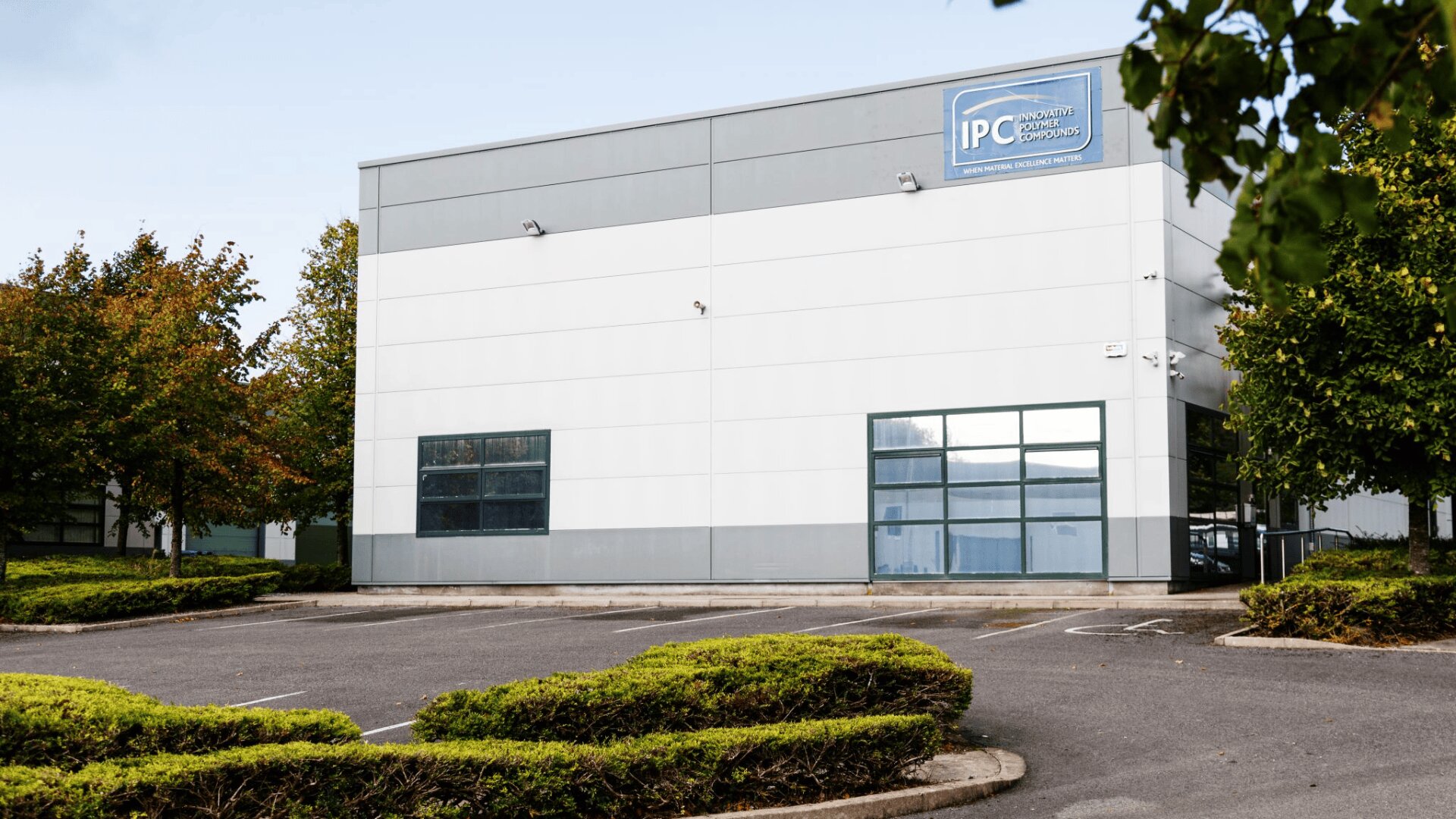 A modern, white and gray office building with large windows, a blue IPC sign at the top, and an empty parking lot surrounded by greenery and trees.