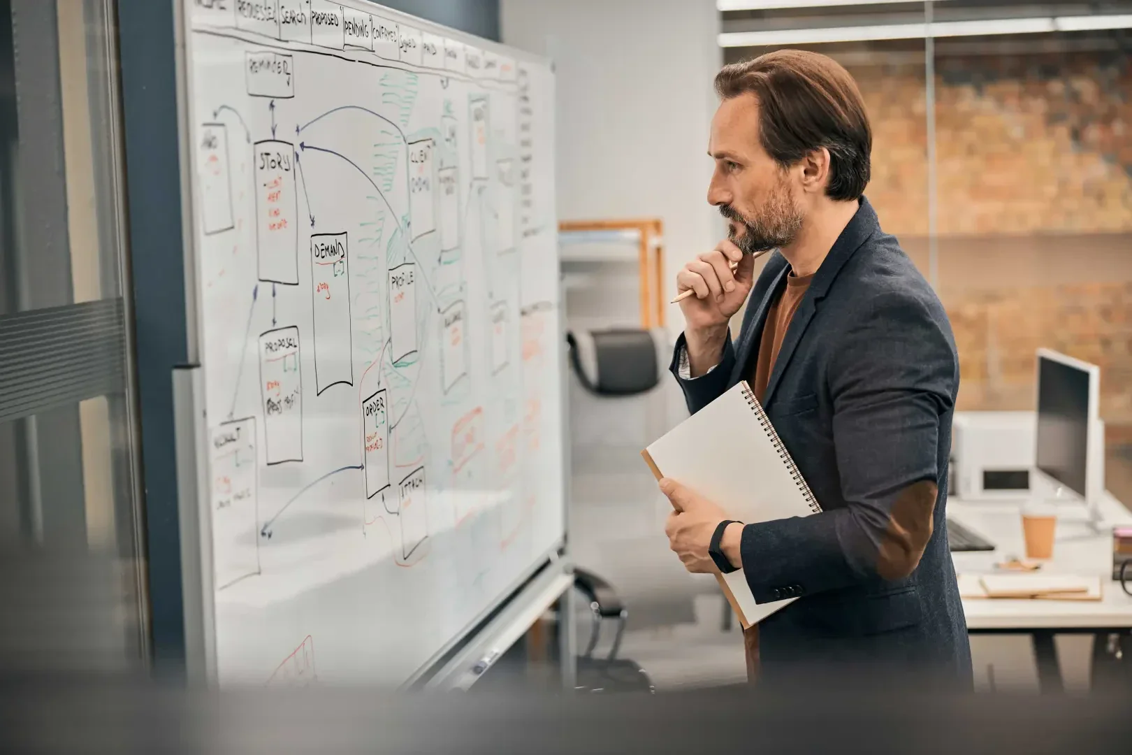 A man in a suit holds a notebook in his hand and looks thoughtfully at a board with diagrams, tables and notes in an office.