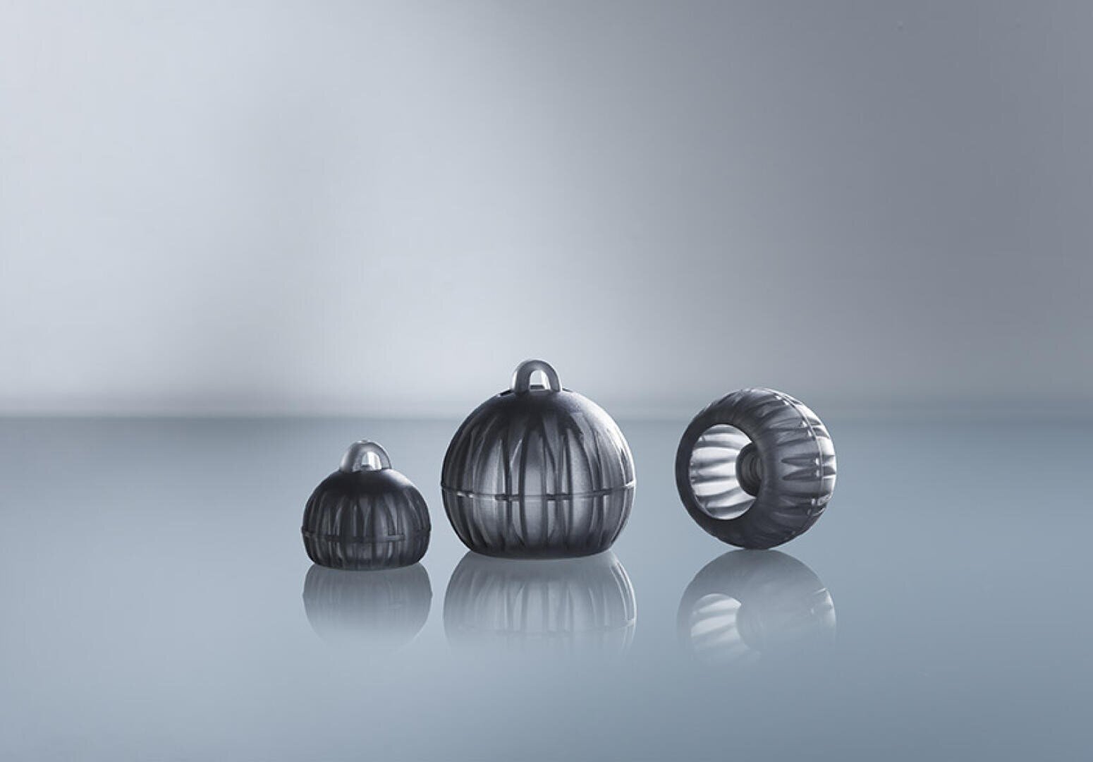Three spherical, metallic objects with vertical grooves are displayed on a reflective surface. Two stand upright, one larger than the other, and the third lies on its side, revealing a hollow interior. The background is soft and has a color gradient.