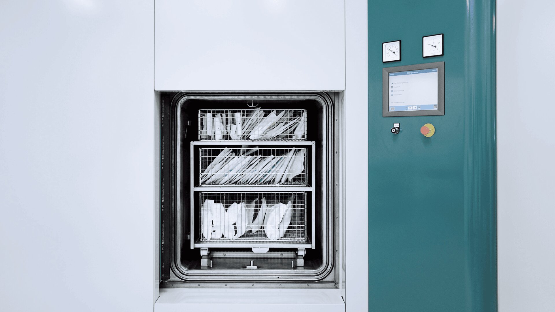 Eine große Sterilisationsmaschine mit geöffneter Tür und Metallgestellen, die mit medizinischen Instrumenten gefüllt sind. Auf der rechten Seite der Maschine ist das Bedienfeld mit Messgeräten und einem digitalen Bildschirm zu sehen.
