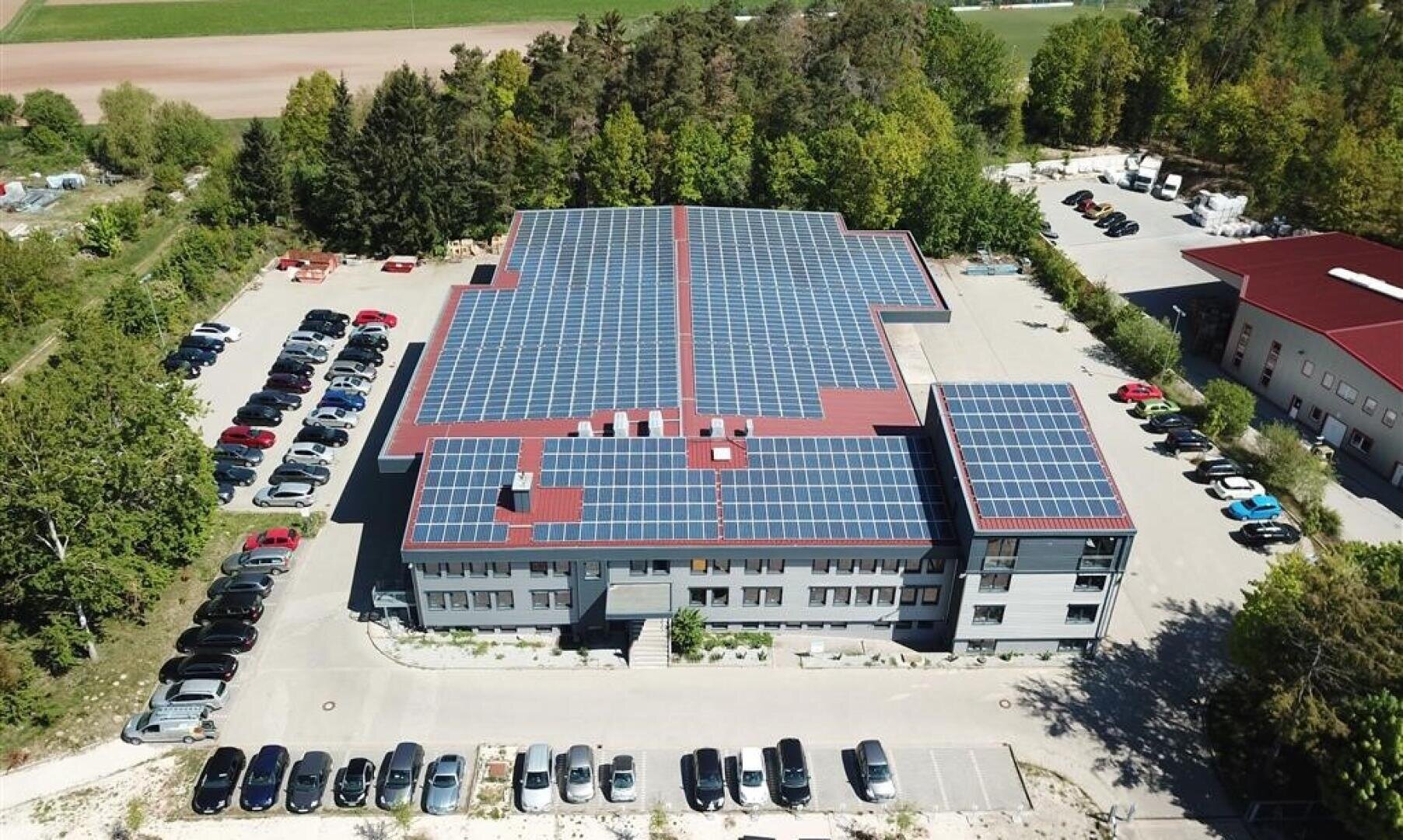 Luftaufnahme eines großen Gebäudes mit Sonnenkollektoren, die den größten Teil des Daches bedecken, umgeben von geparkten Autos und Bäumen. Die Umgebung scheint halb-ländlich zu sein, mit offenen Feldern und bewaldeten Gebieten in der Nähe.