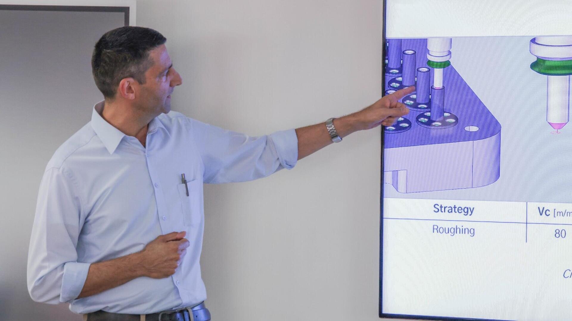 A man in a light blue shirt points at a digital screen displaying a technical diagram with the word Strategy and numerical values, appearing to explain or present engineering data.