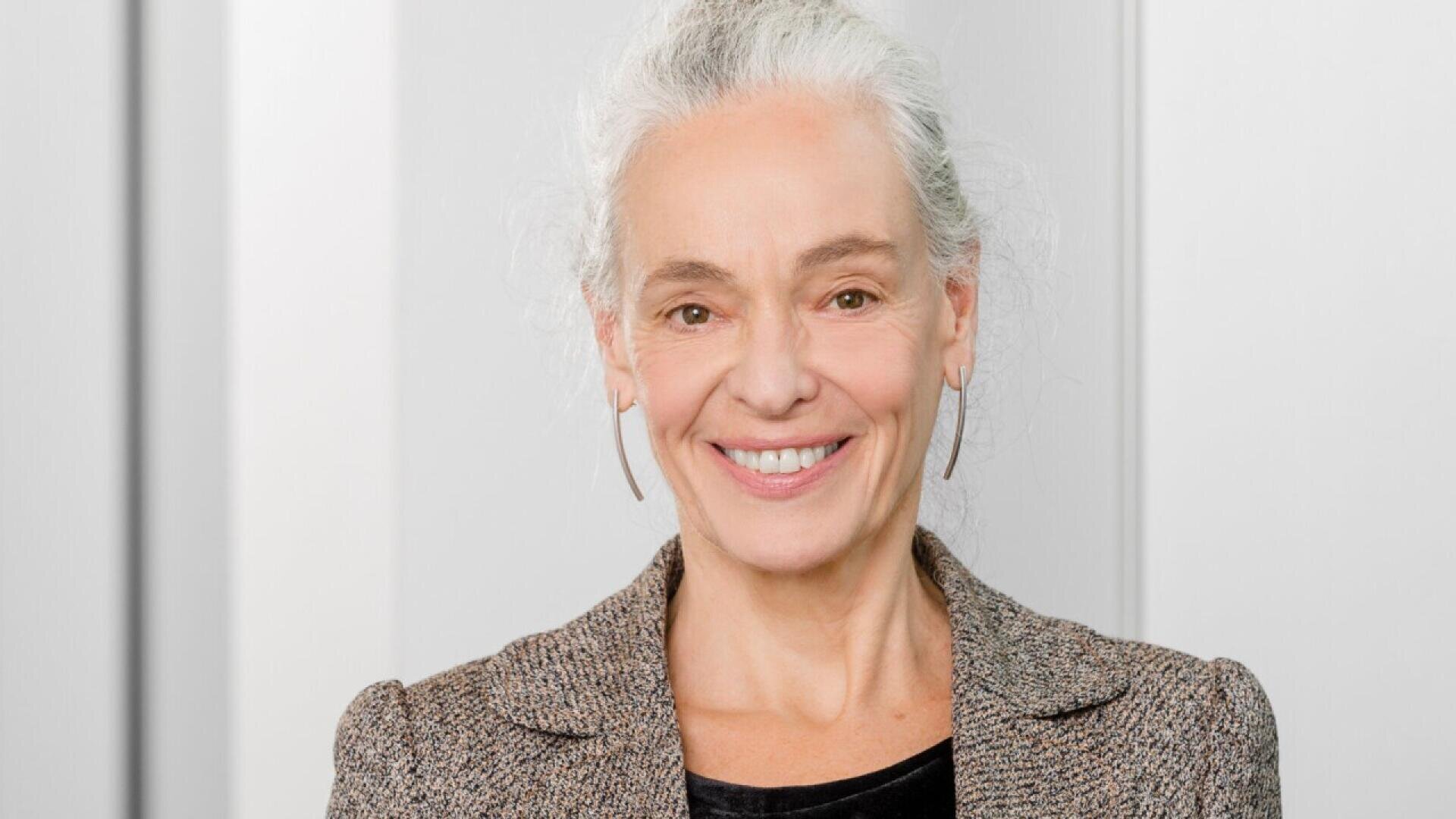 A smiling older woman with gray hair pulled back, wearing a brown textured blazer and hoop earrings, stands in front of a light-colored background.