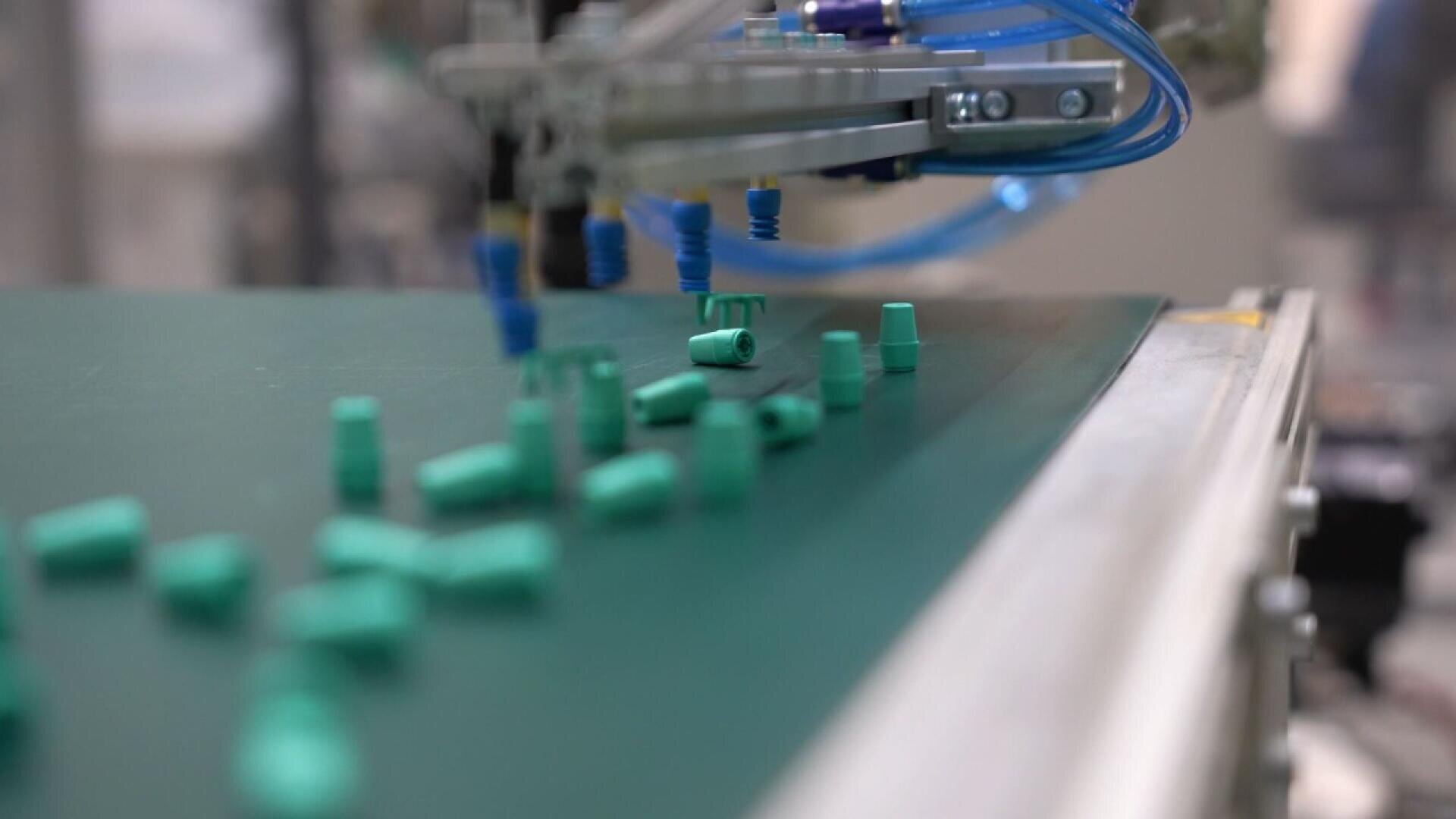 A close-up of a factory conveyor belt with green plastic components and a robotic arm with blue suction cups sorting the parts. The background is blurred, focusing on the automation process.