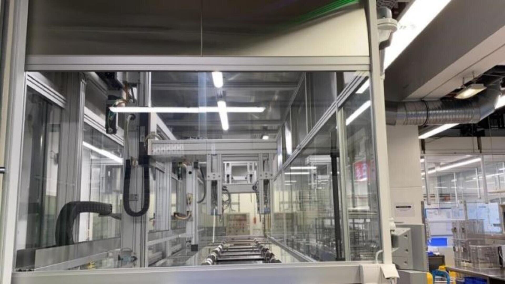 A modern factory interior with automated machinery, glass panels, metal frames, and conveyor belts under bright overhead lighting. Industrial equipment and pipes are visible in the background.