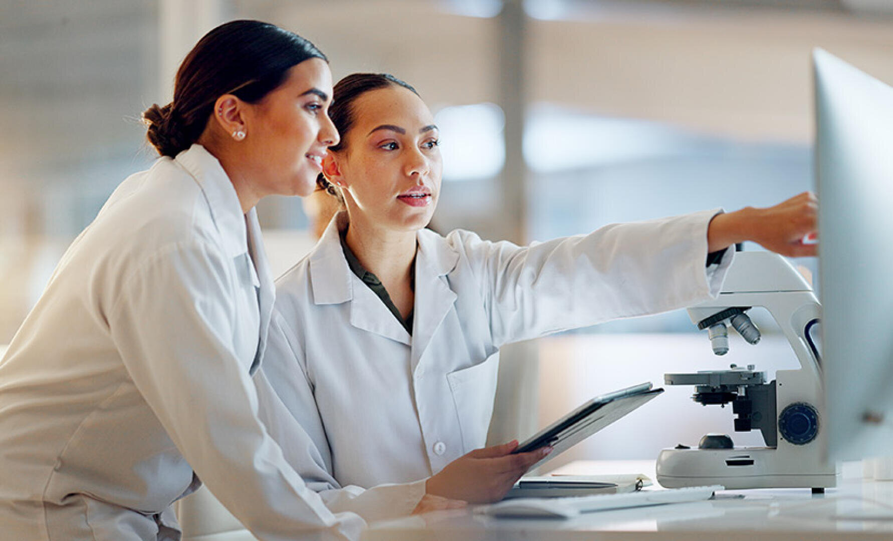 Two women in lab coats work together in a laboratory. One points to a computer screen while the other holds a tablet. A microscope is visible on the desk next to them.