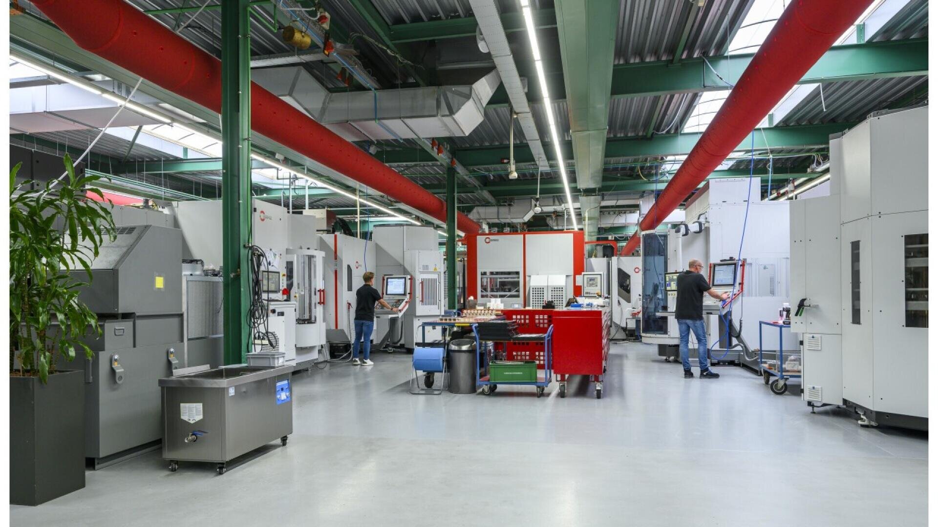 A modern industrial workshop with several large machines, computer terminals, and two people operating equipment. The space is brightly lit with overhead lighting and exposed ducts. Tools and carts are scattered throughout the area.