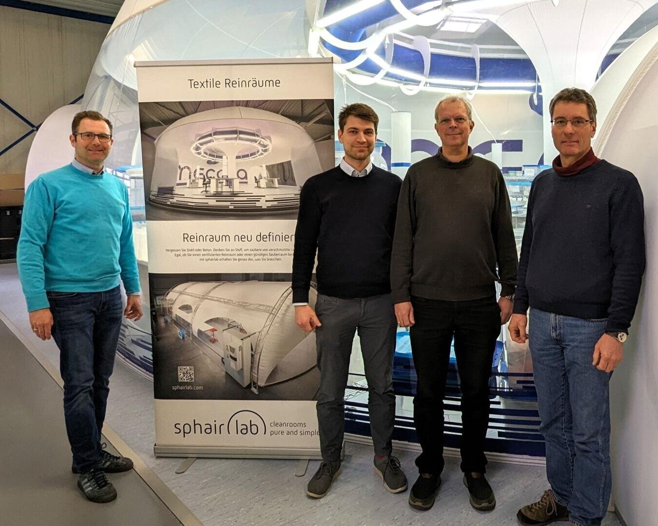 Vier Männer stehen im Innenbereich neben einem großen vertikalen Banner von sphairlab, das Bilder und Texte über textile Reinräume zeigt. Die Umgebung erinnert an einen modernen Industrie- oder Laborraum.