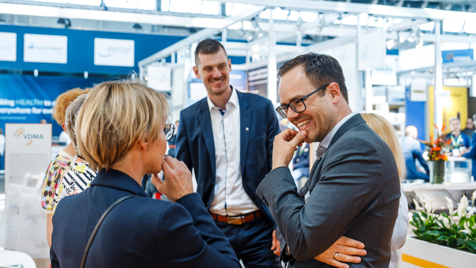 Eine Gruppe professionell gekleideter Menschen steht lächelnd in einer hellen Ausstellungshalle und unterhält sich freundlich in der Nähe von Ausstellungsständen.