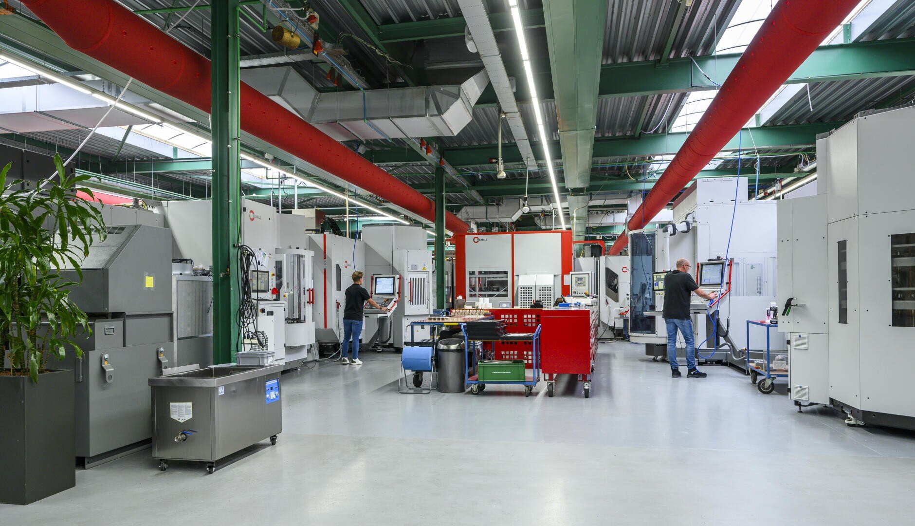 Eine geräumige, moderne Fabrikhalle mit mehreren großen Industriemaschinen. Arbeiter bedienen Geräte, Monitore und Steuerungen. An der Decke sind Rohre und Beleuchtung freigelegt, und überall stehen Karren und Werkzeugkästen.