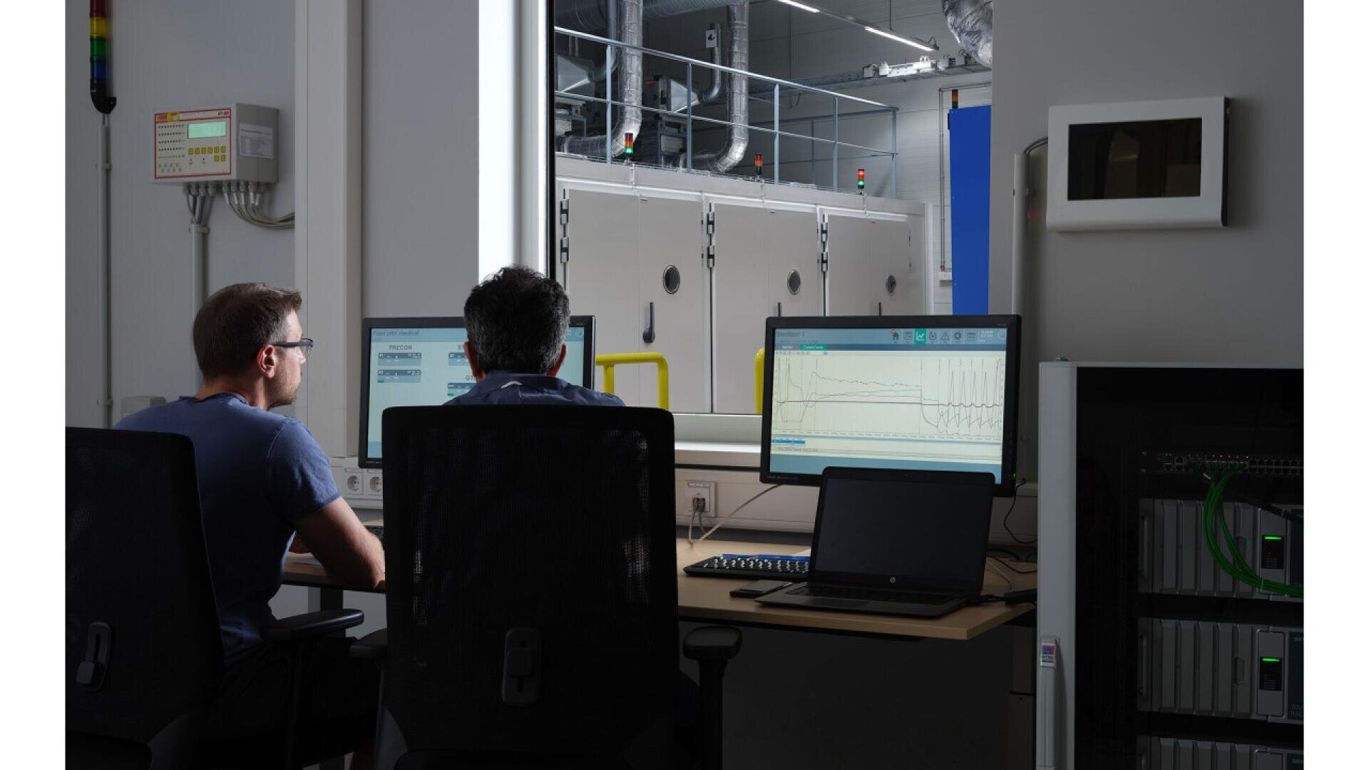 Two people sit at desks with computer monitors displaying graphs and data, facing a large window that looks into an industrial lab with machinery and equipment. The room is dimly lit, focusing on the workstations.