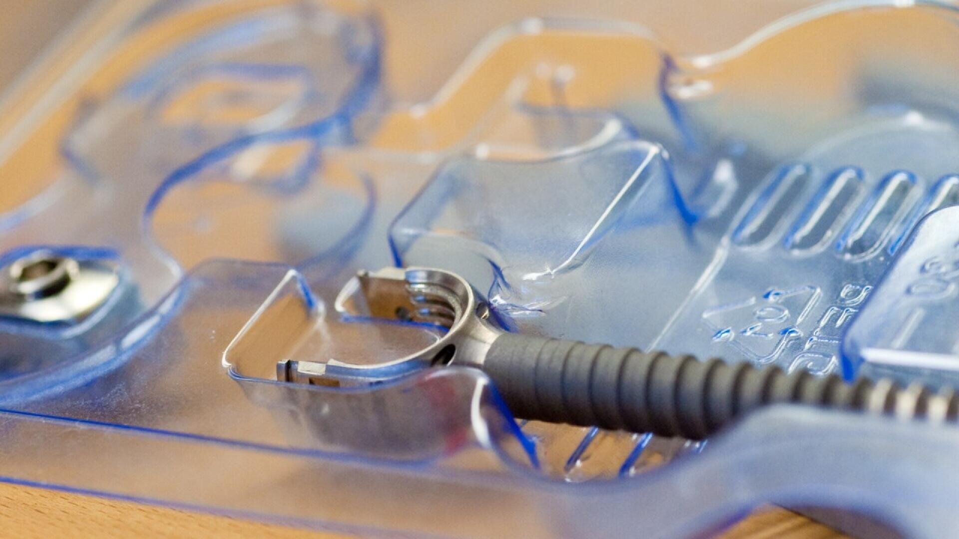 A close-up of a dental instrument inside a transparent blue plastic packaging tray, resting on a wooden surface. The tool has a metallic tip and a textured handle.