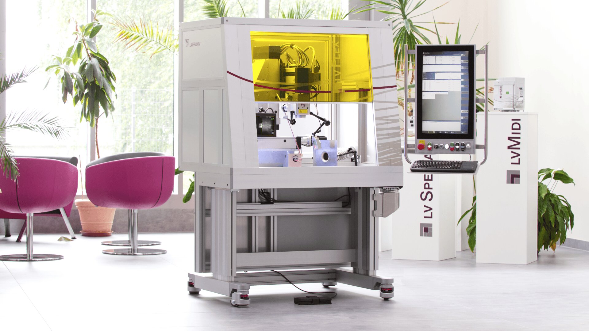 Eine Hightech-Labormaschine mit gelb getönter Schutzhülle steht in einem modernen, hellen Raum mit Bedienfeld, Topfpflanzen und violetten Stühlen im Hintergrund.