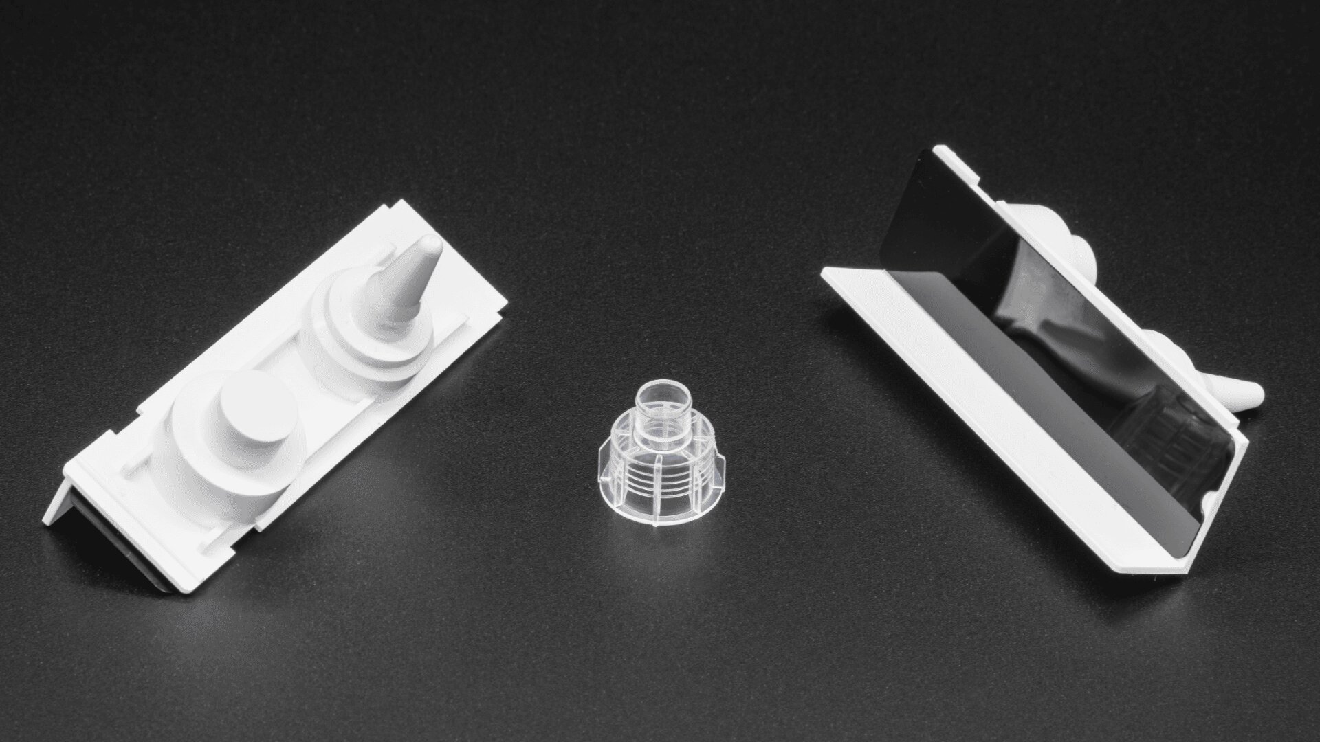 Drei Kunststoffobjekte auf einer schwarzen Oberfläche: ein weißes Stück mit zwei kreisförmigen Formen und einem Kegel auf der linken Seite, ein klares zylindrisches Stück in der Mitte und ein weißes Stück mit einem schwarzen rechteckigen Abschnitt auf der rechten Seite.