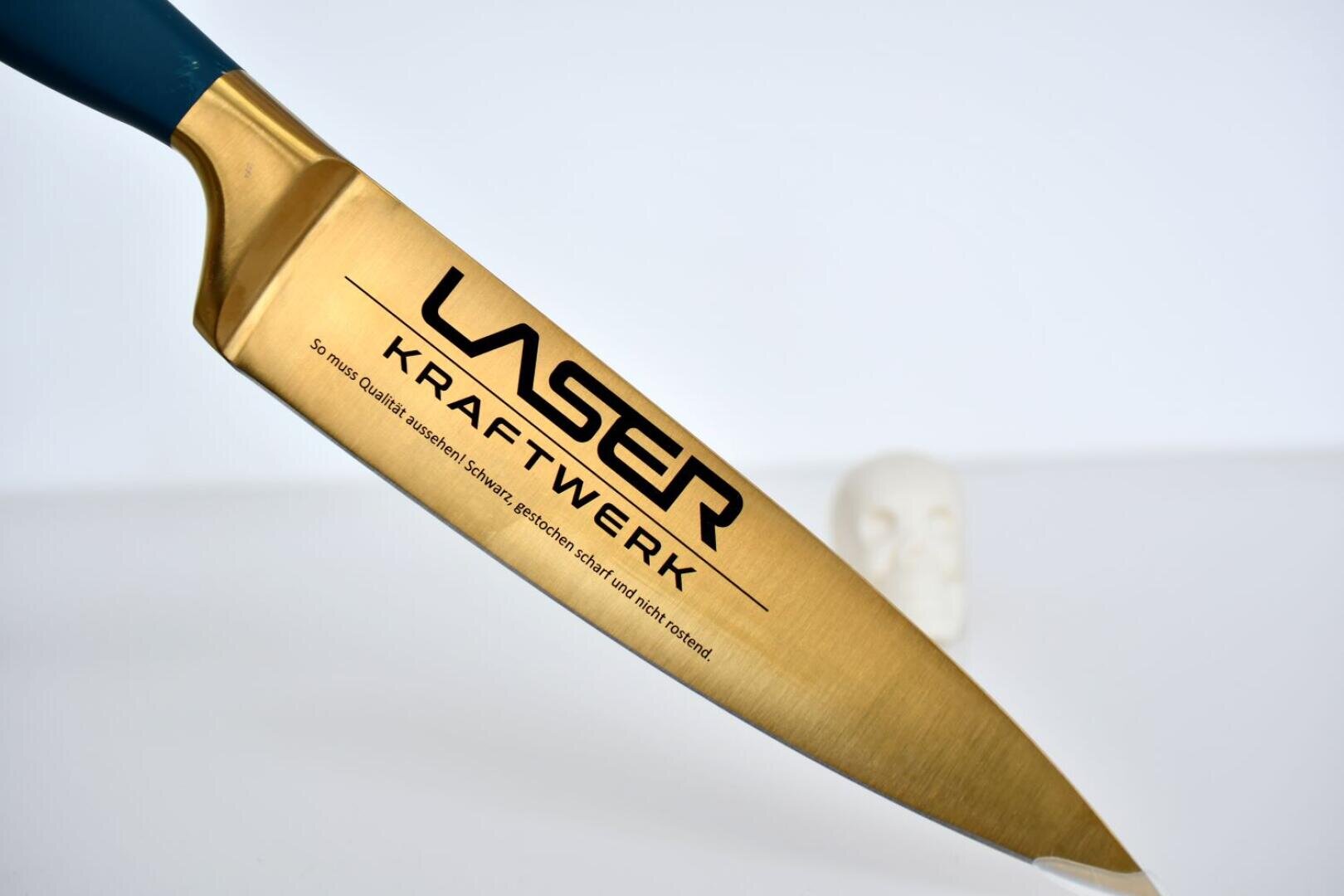 Nahaufnahme einer goldfarbenen Küchenmesserklinge mit aufgedrucktem LASER KRAFTWERK und Produktangaben vor einem weißen Hintergrund mit einem kleinen, verschwommenen weißen Objekt in der Ferne.