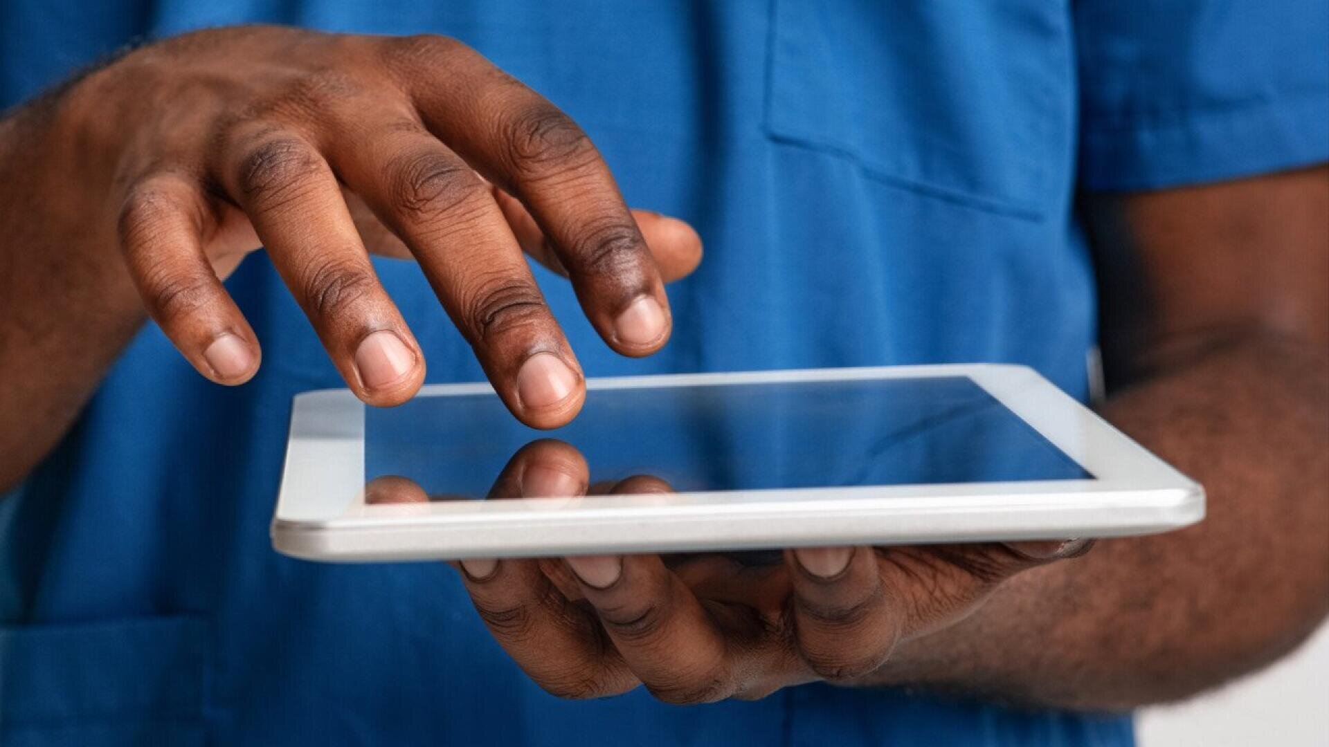 A person wearing blue scrubs uses a tablet, tapping the screen with one hand while holding the device with the other. The focus is on the hands and the tablet.