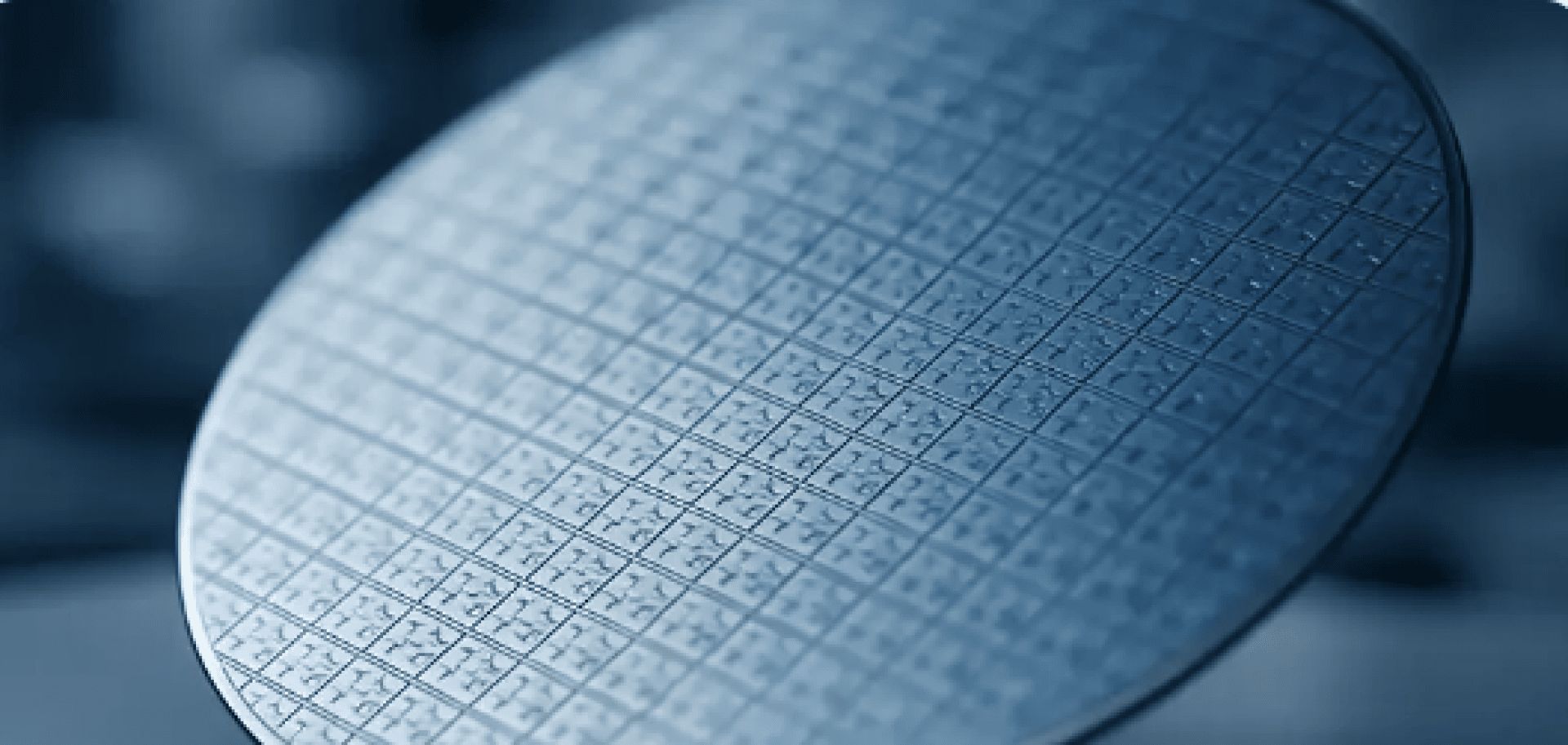 Close-up of a silicon wafer covered with a grid pattern of small microchips, reflecting blue light in a high-tech production environment.