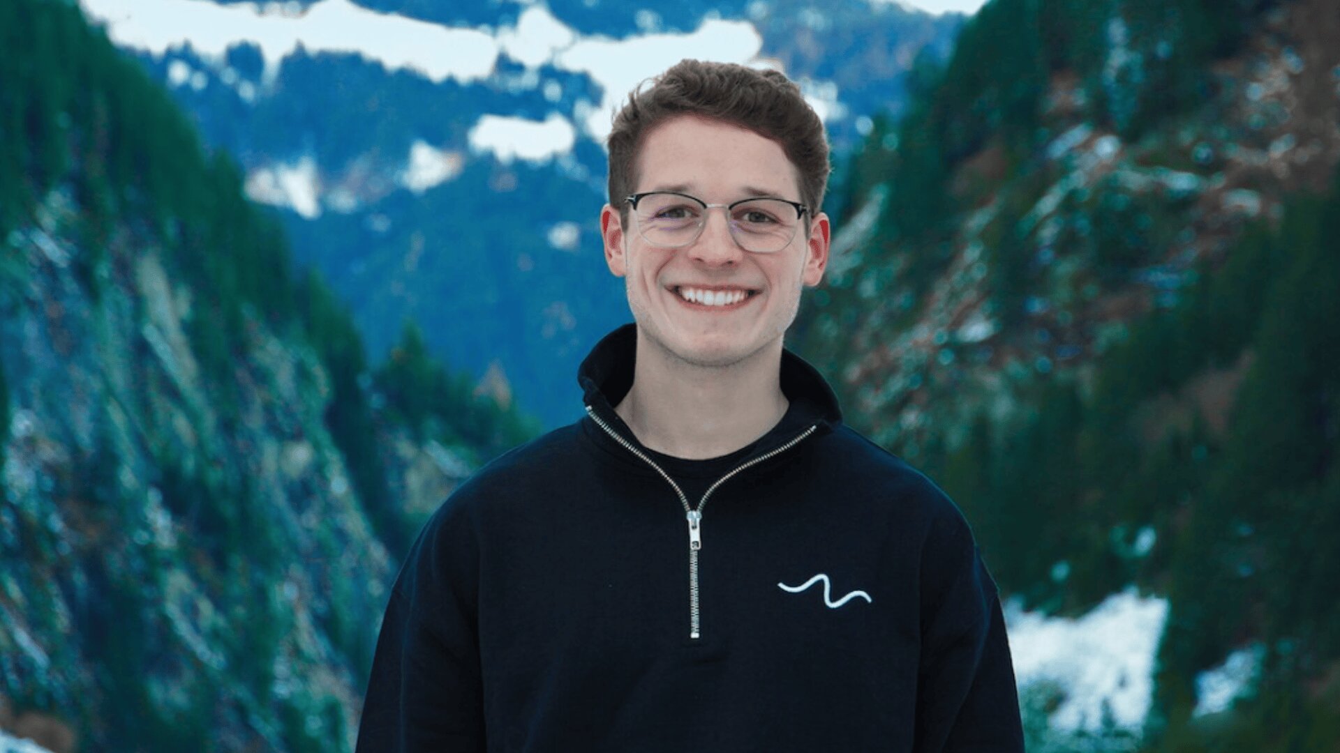 A young man wearing glasses and a black zip-up sweater stands smiling outdoors with snowy mountains and pine trees in the background.
