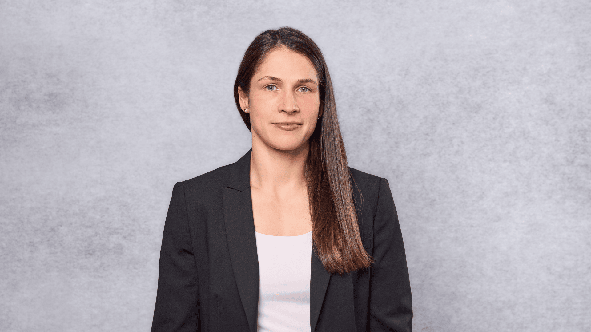 Eine Frau mit glattem braunem Haar, die einen schwarzen Blazer und ein helles Oberteil trägt, steht vor einem einfarbigen, hellgrau strukturierten Hintergrund und blickt mit neutralem Blick direkt in die Kamera.