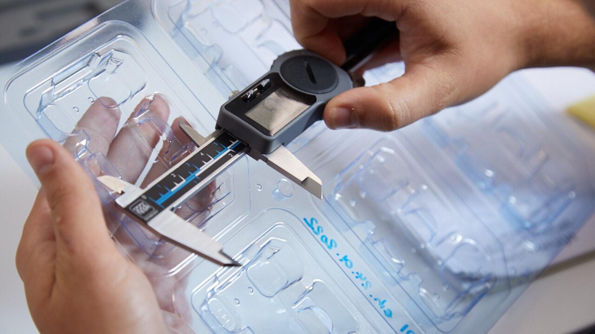 A person uses a digital caliper to measure the thickness of a clear plastic packaging tray, holding the tray in one hand for precise measurement.