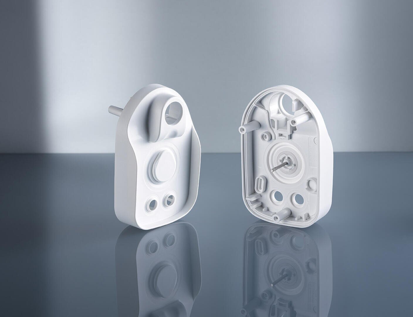 Two white plastic electronic device enclosures stand upright on a glossy surface, one showing the smooth outer shell and the other displaying the detailed inner components and mounting features.
