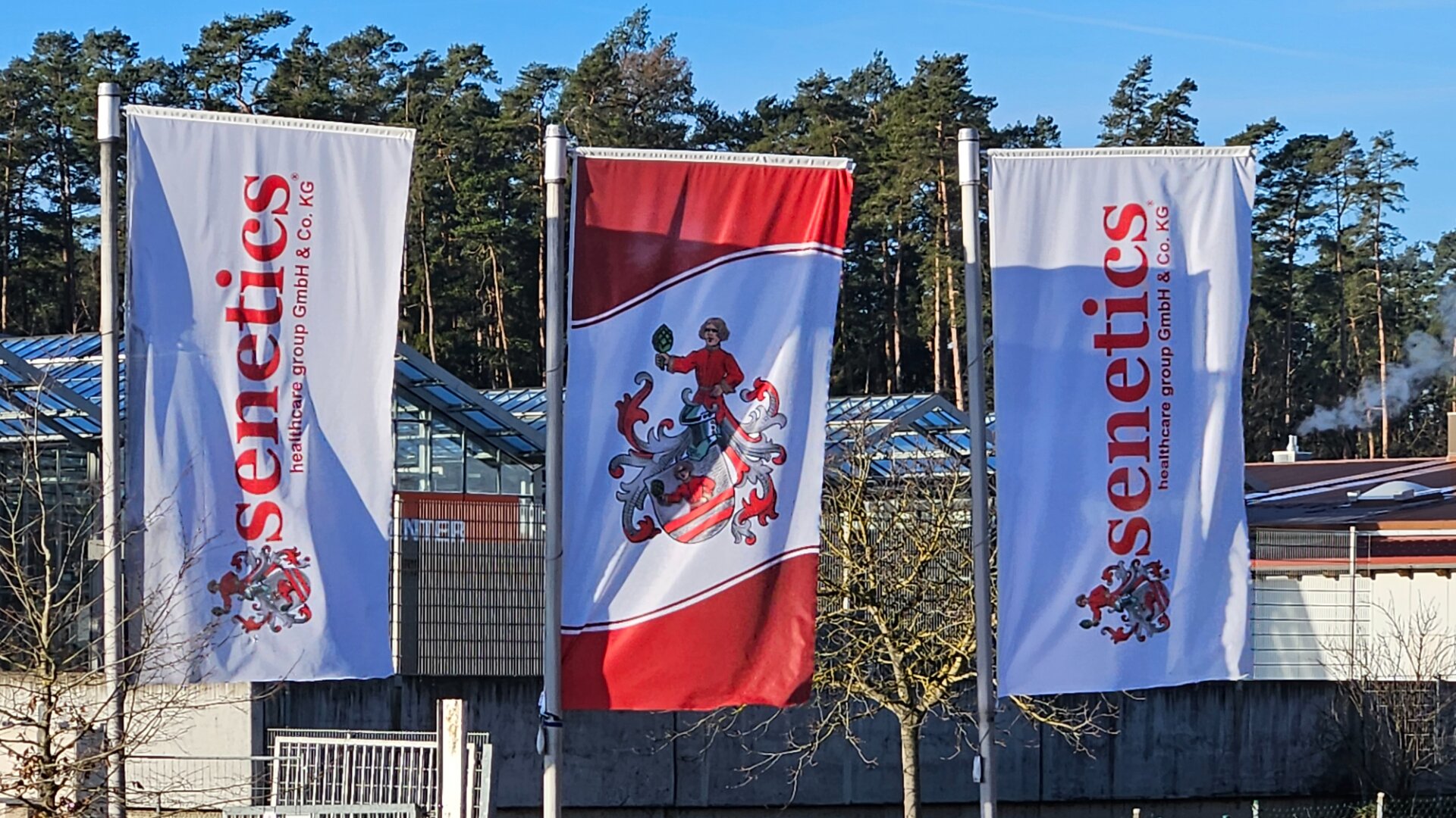 Draußen hängen drei Fahnen an Masten, zwei mit Senetics und eine in der Mitte mit einem rot-weißen Wappen mit einem Ritter. Im Hintergrund sind Bäume, ein Gewächshaus und ein Gebäude zu sehen.