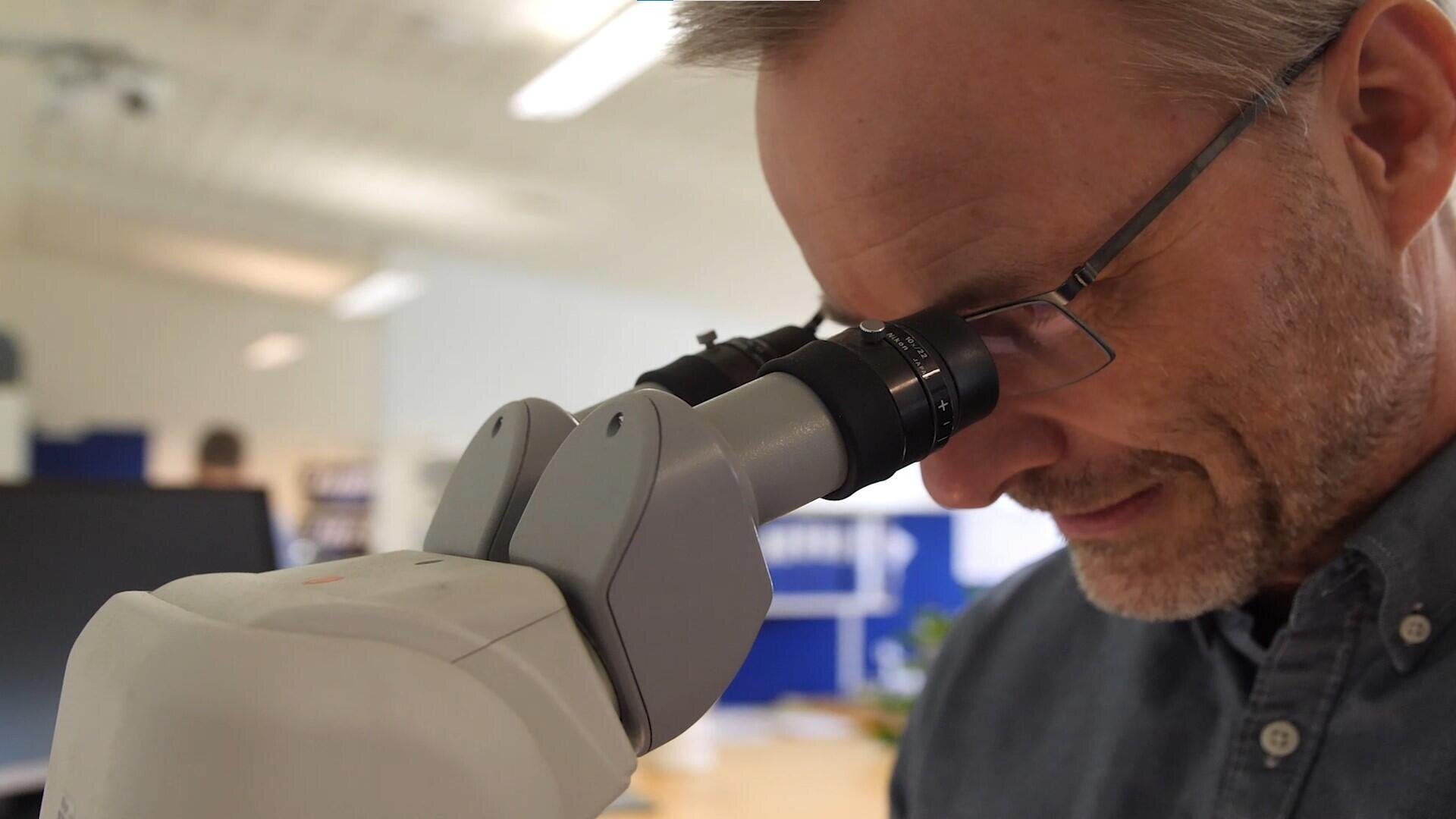 A man with glasses examines something through a microscope in a bright interior of a laboratory or office.