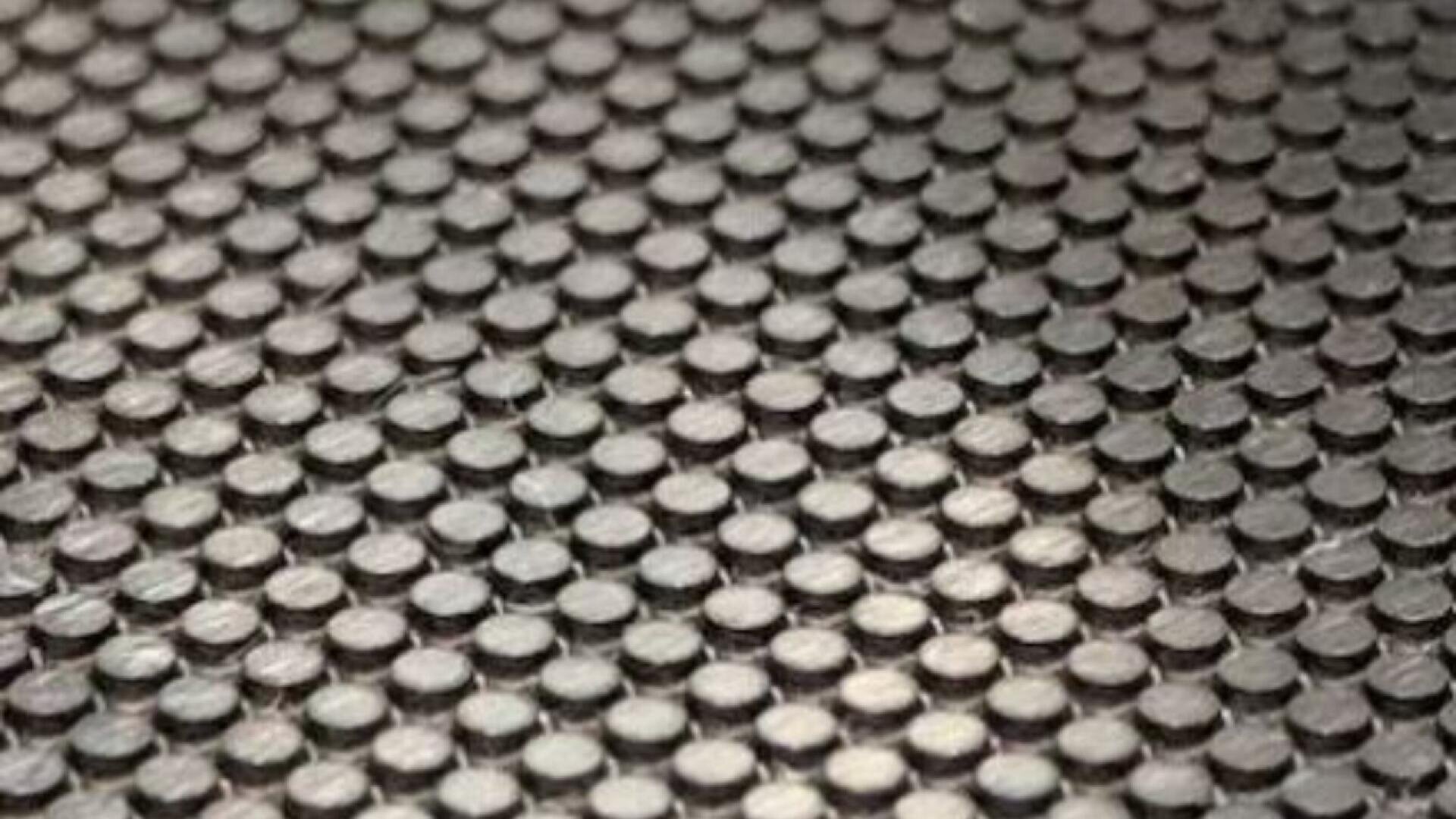 Close-up of a textured surface featuring a repeating pattern of small, round, raised bumps arranged in a grid. The material appears metallic or plastic, with a matte finish and subtle reflections.