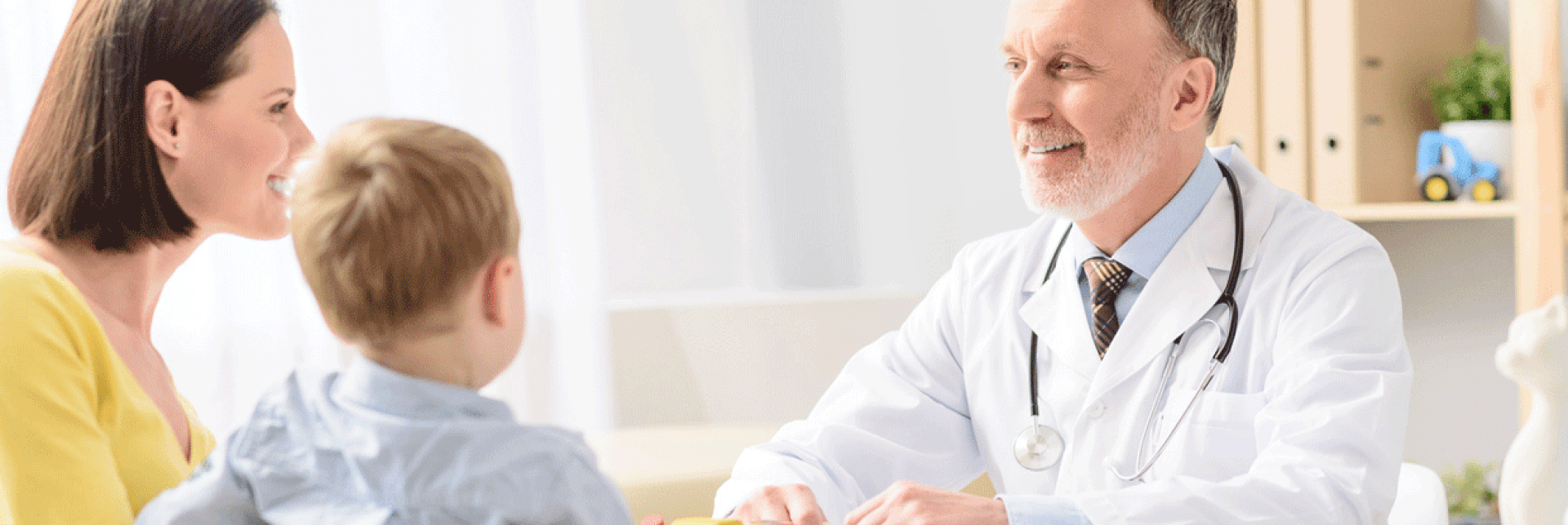 A smiling doctor in a white coat sits at a desk and speaks with a woman and a boy sitting across from him in a bright office.