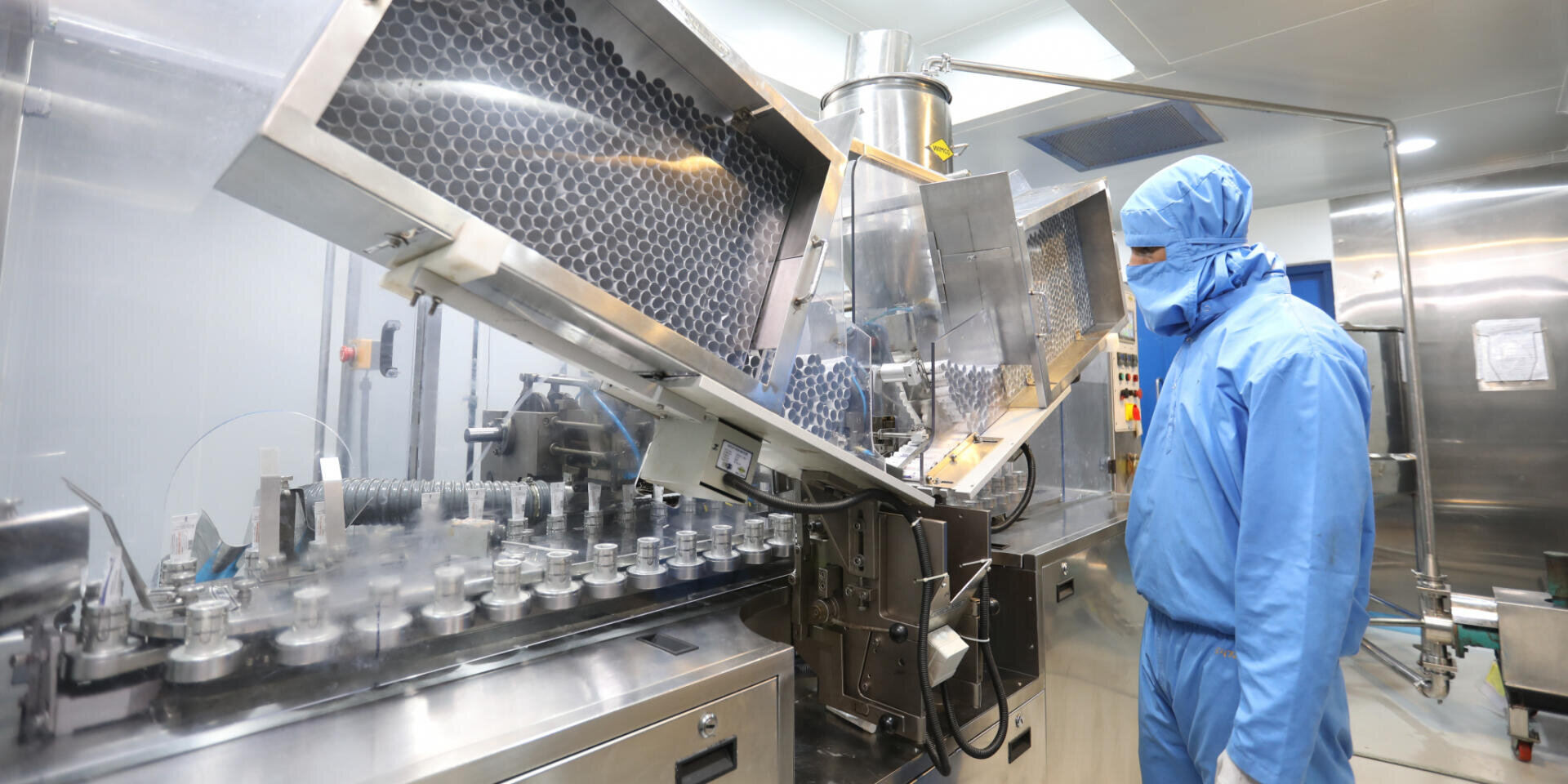 Eine Person in blauer Schutzkleidung steht in einem Reinraum und überwacht automatisierte Maschinen, die Fläschchen in einer pharmazeutischen oder Laborumgebung abfüllen. Edelstahlgeräte und sterile Bedingungen sind sichtbar.