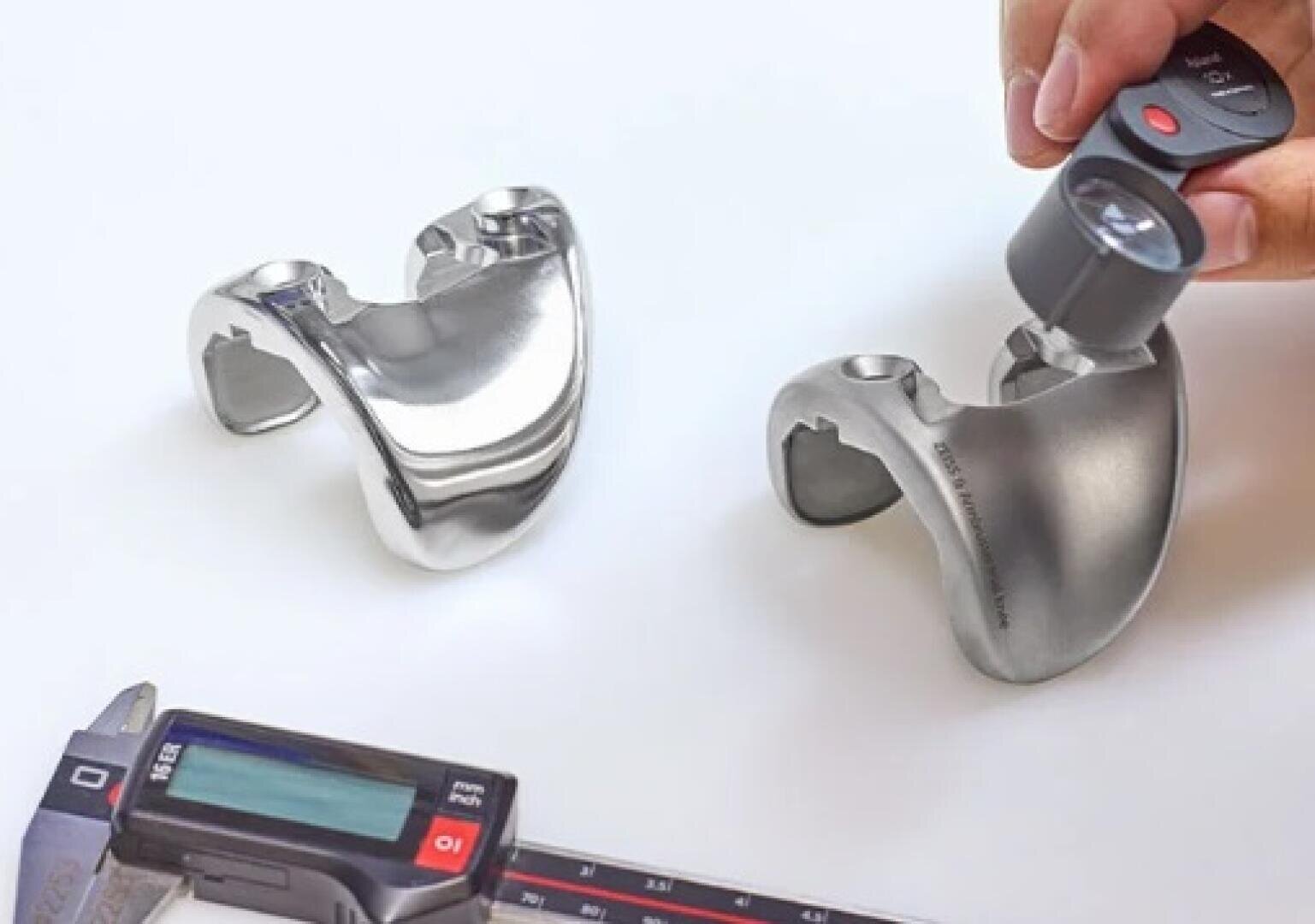 Zwei metallisch glänzende Knieimplantate sind auf einer weißen Fläche platziert. In der Nähe befinden sich ein Messschieber und eine Lupe, die von einer Hand gehalten werden, was darauf hindeutet, dass die Teile auf Qualität oder Präzision geprüft werden.