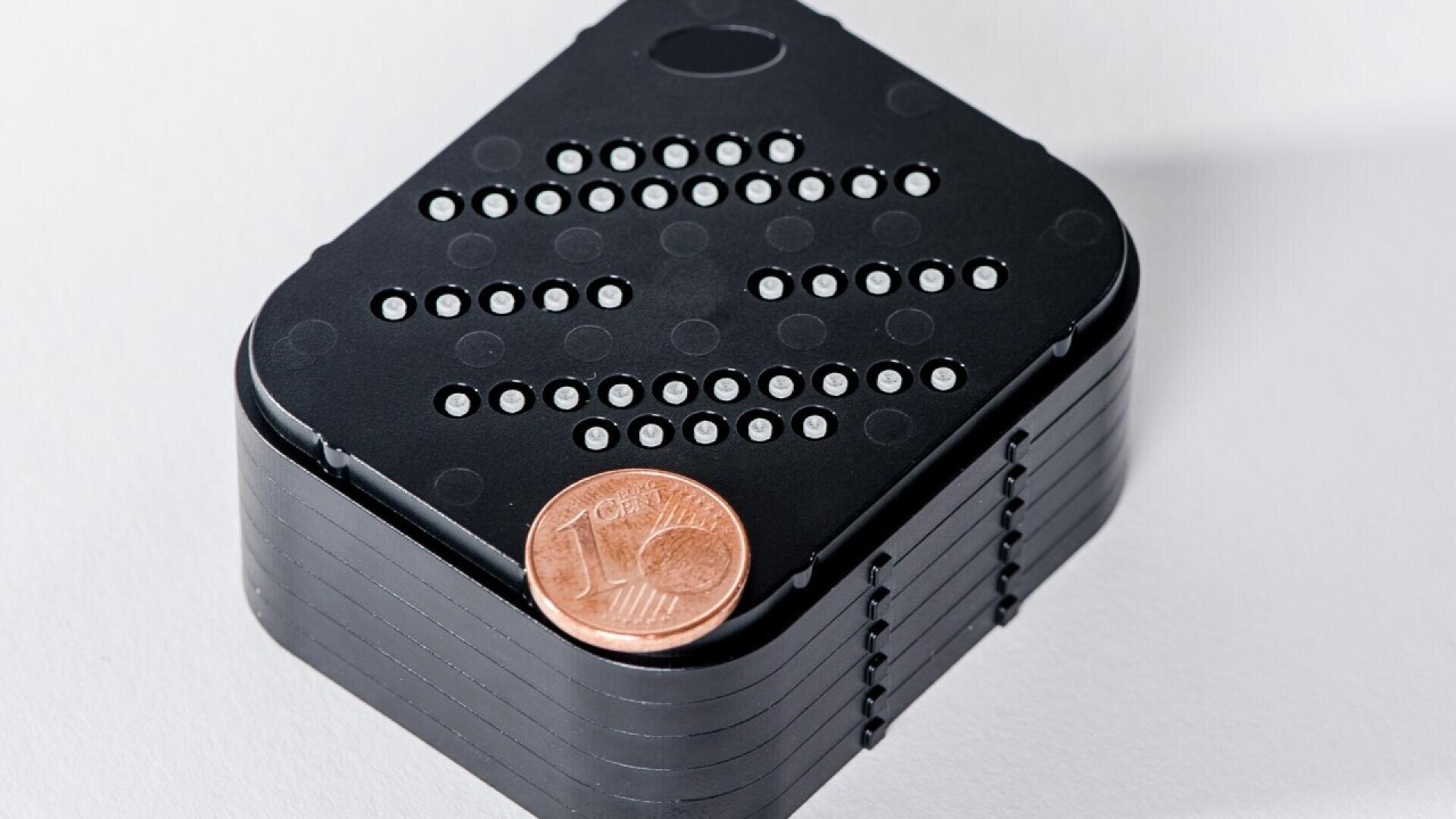 A small black rectangular device with circular holes on its top, stacked in layers, is shown with a 10 cent euro coin placed on its corner for scale. The background is plain white.