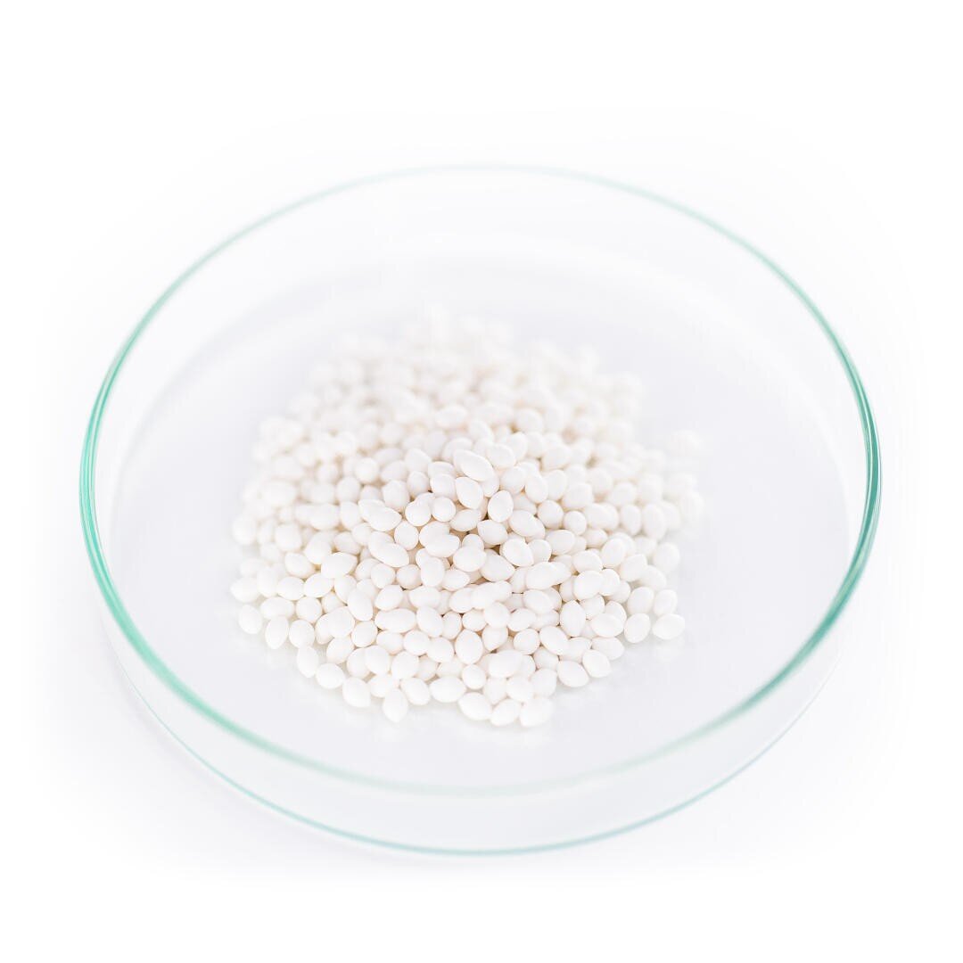 A clear glass petri dish filled with small, round, white pellets is shown on a white background.