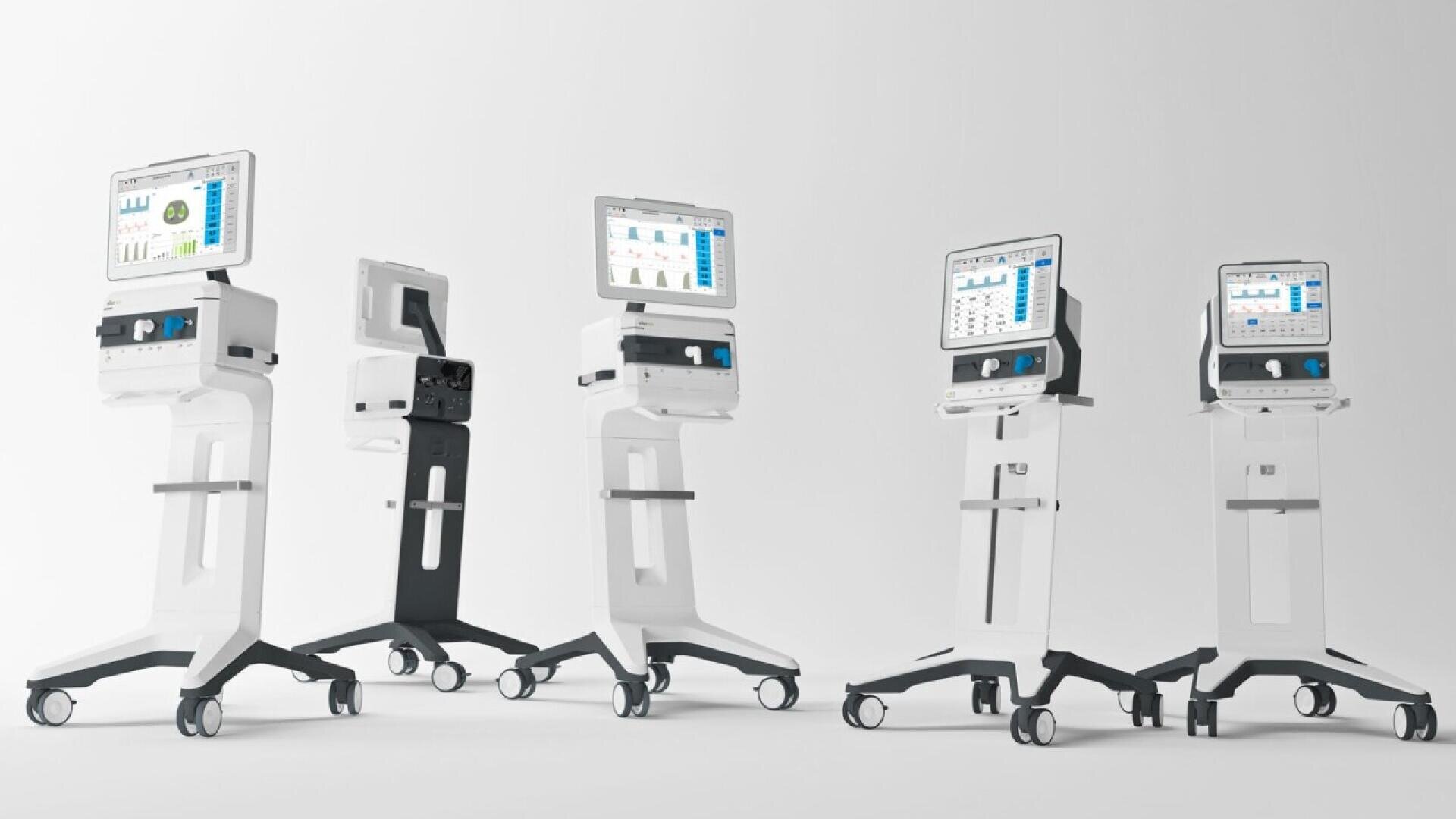 Four modern medical ventilators on wheels, each with screens displaying graphs and data, are arranged in a row against a plain white background. The ventilators have sleek, white designs with dark accents.